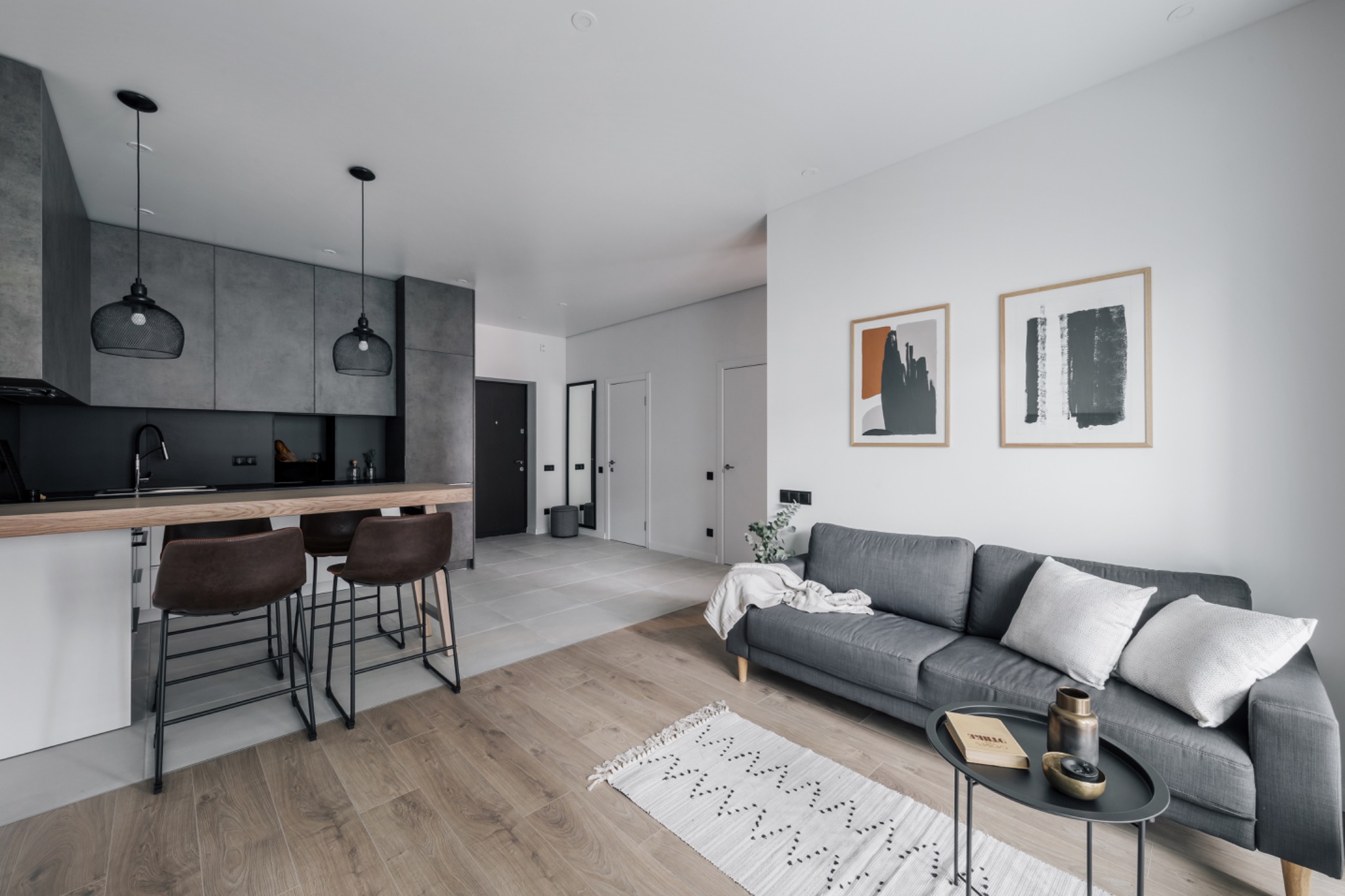 Nowoczesne wnętrze mieszkania w odcieniach szarości i brązu, z drewnianą podłogą, szarą sofą z poduszkami, stolikiem kawowym z dekoracjami, otwartą kuchnią z wyspą i hokerami oraz dywanikiem...
