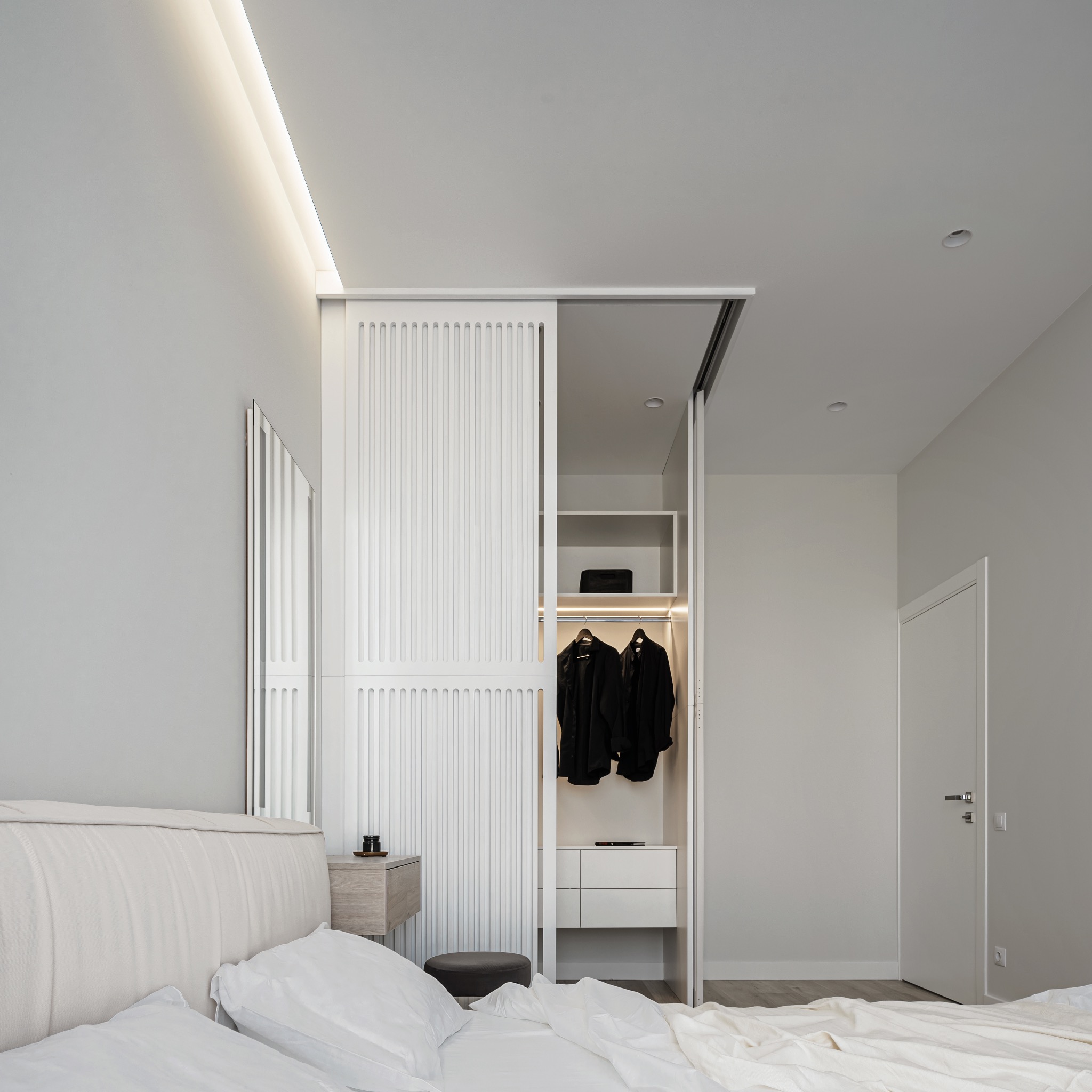Minimalistyczna sypialnia z łóżkiem z pikowanym zagłówkiem i szafą z przesuwnymi lamelowymi drzwiami, oświetloną od wewnątrz.