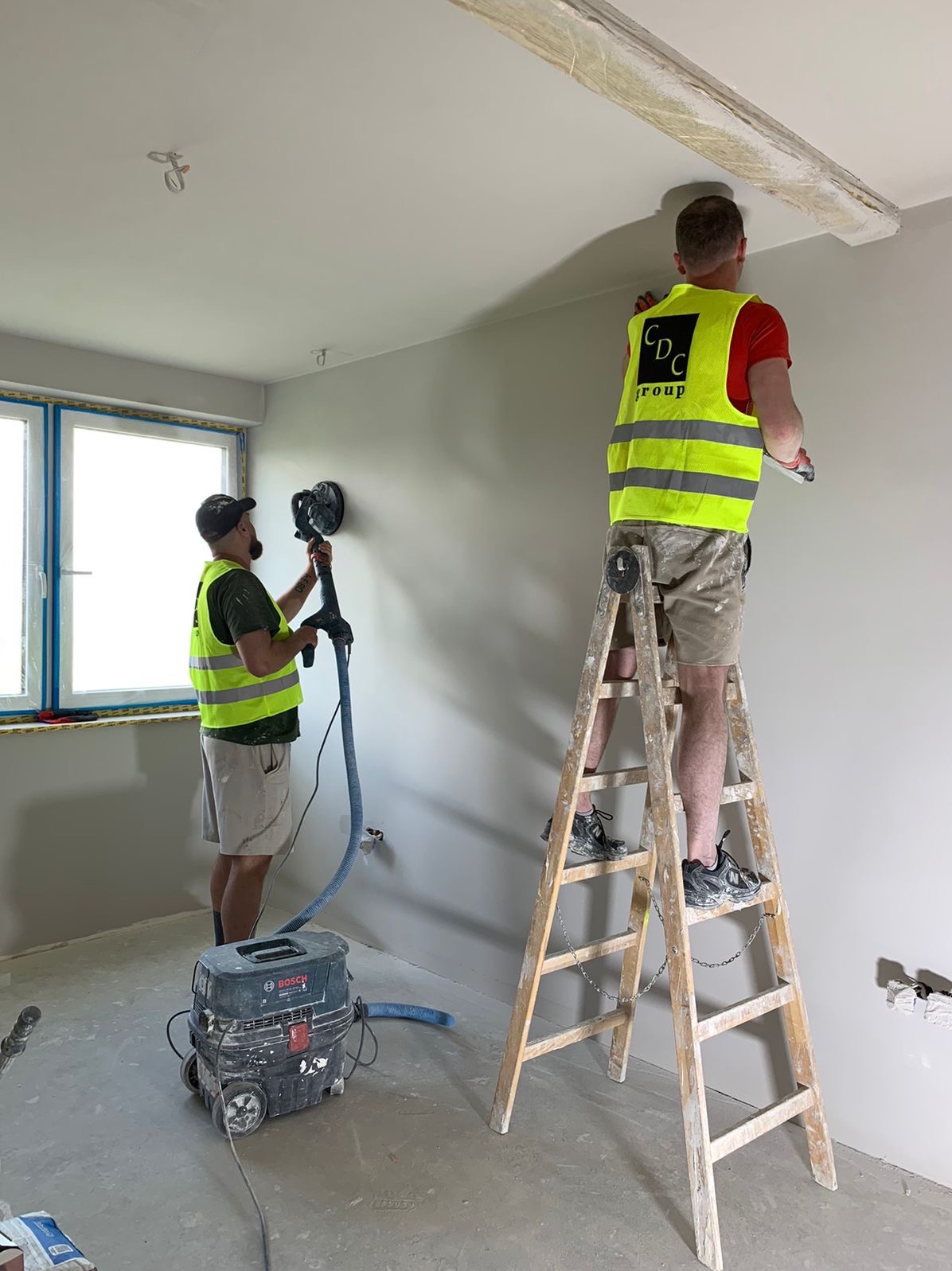 Dwóch pracowników w odblaskowych kamizelkach szlifuje ściany i sufit w nowym pomieszczeniu, używając szlifierki z odkurzaczem Bosch, jeden na drabinie.