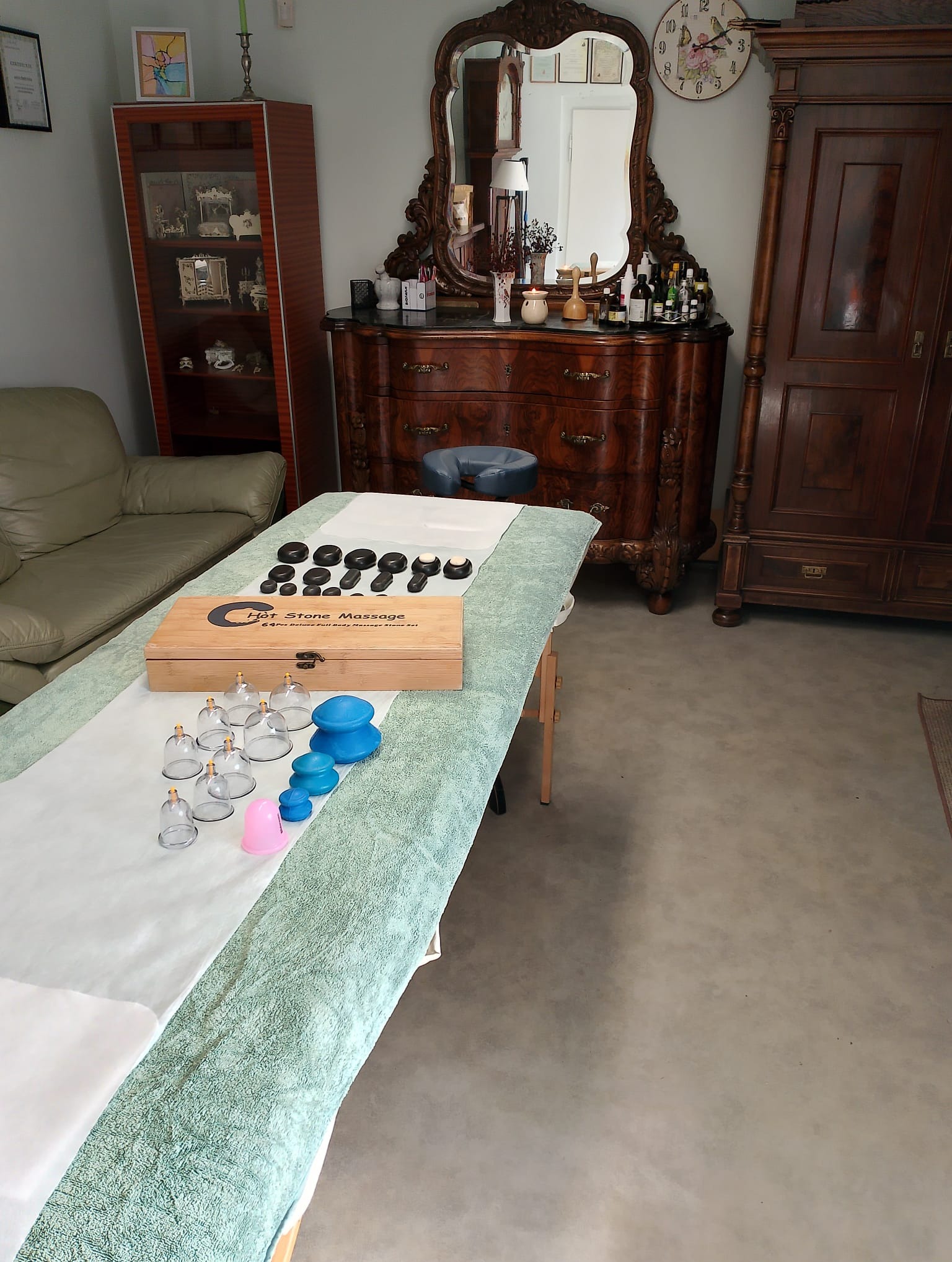 Gabinet z kozetką, zestawem do masażu gorącymi kamieniami w drewnianym pudełku, bańkami szklanymi i silikonowymi w odcieniach niebieskiego i różowego, komodą z lustrem i zegarem oraz witryną.