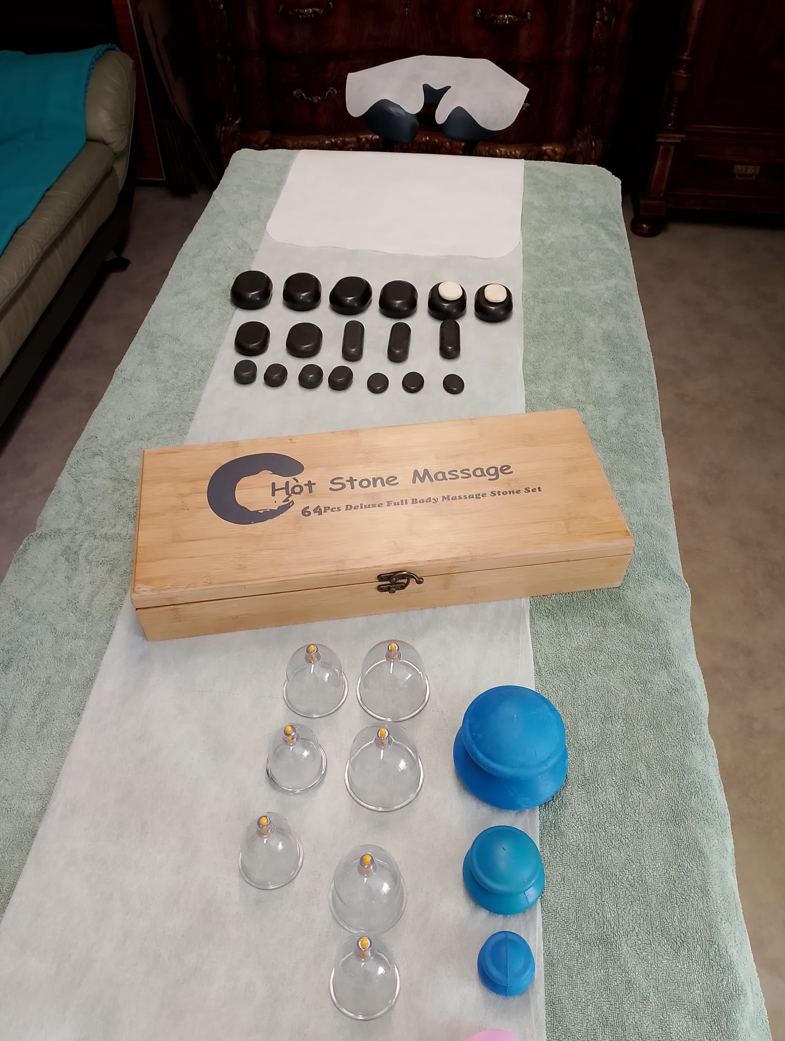Przygotowany stół do zabiegu z kamieniami i bańkami: drewniane pudełko z napisem 'Hot Stone Massage', zestaw czarnych kamieni do masażu, szklane i niebieskie bańki do terapii próżniowej, całość...