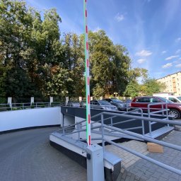 POTEREK AUTOMATIC - DAMIAN POTEREK - Szlaban automatyczny pionowy, biało-czerwony, z zielonym oświetleniem LED, przy wjeździe do garażu podziemnego, z widocznymi samochodami i drzewami w tle.