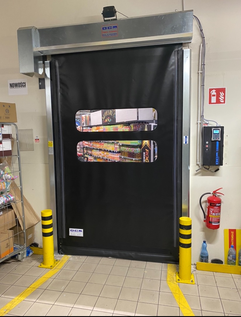 Czarna, szybka brama z dwoma okienkami w magazynie sklepu, widoczne regały z towarem, żółte słupki ochronne i oznakowanie podłogi.