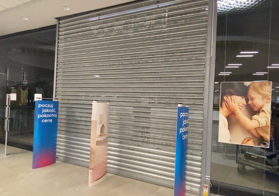 Zasunięta metalowa krata w sklepie z reklamowymi standami po bokach, widok z wnętrza centrum handlowego.