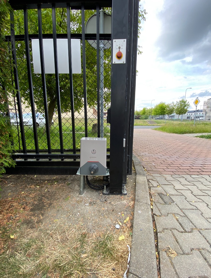 Czarna brama przemysłowa z zamontowanym szarym napędem elektrycznym i przyciskiem awaryjnym, widok z dołu pod lekkim kątem, na tle chodnika z kostki i zieleni.