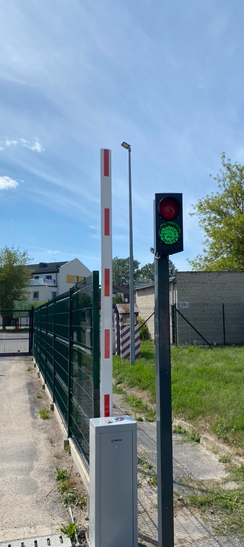 Automatyczna bariera drogowa z sygnalizacją świetlną, widok na osiedle domów jednorodzinnych w tle.