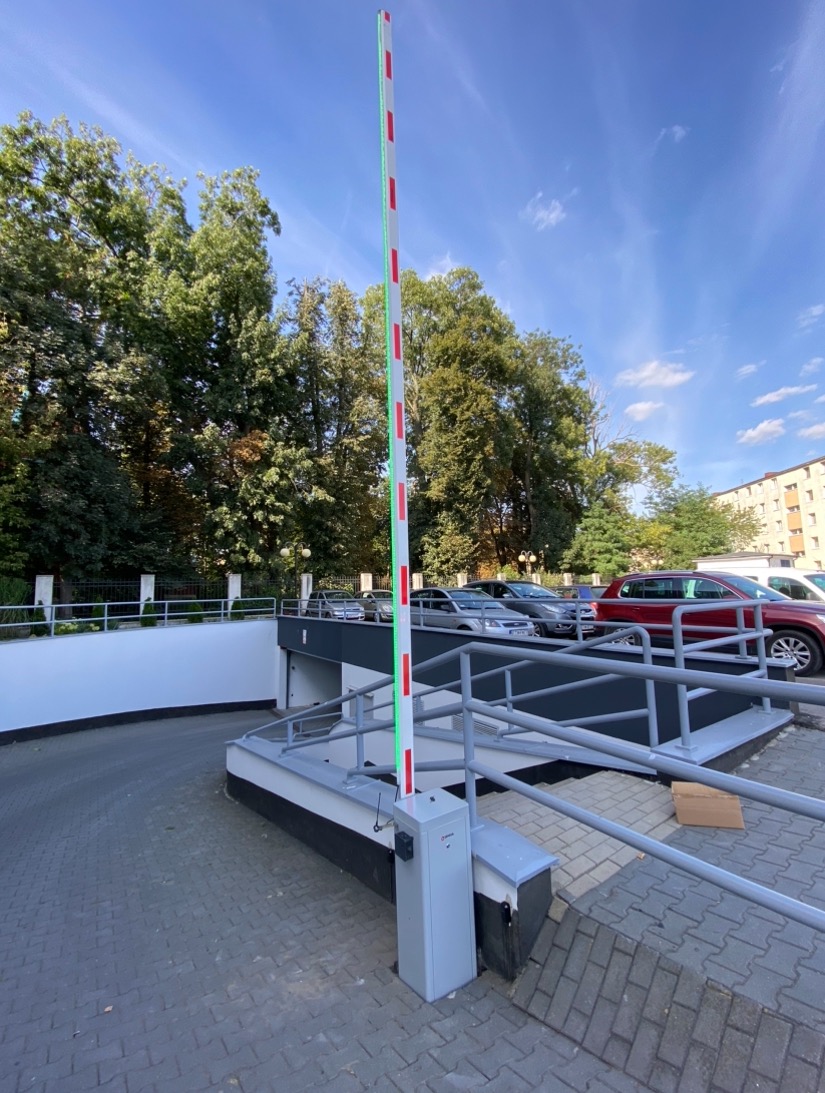 Szlaban automatyczny pionowy, biało-czerwony, z zielonym oświetleniem LED, przy wjeździe do garażu podziemnego, z widocznymi samochodami i drzewami w tle.