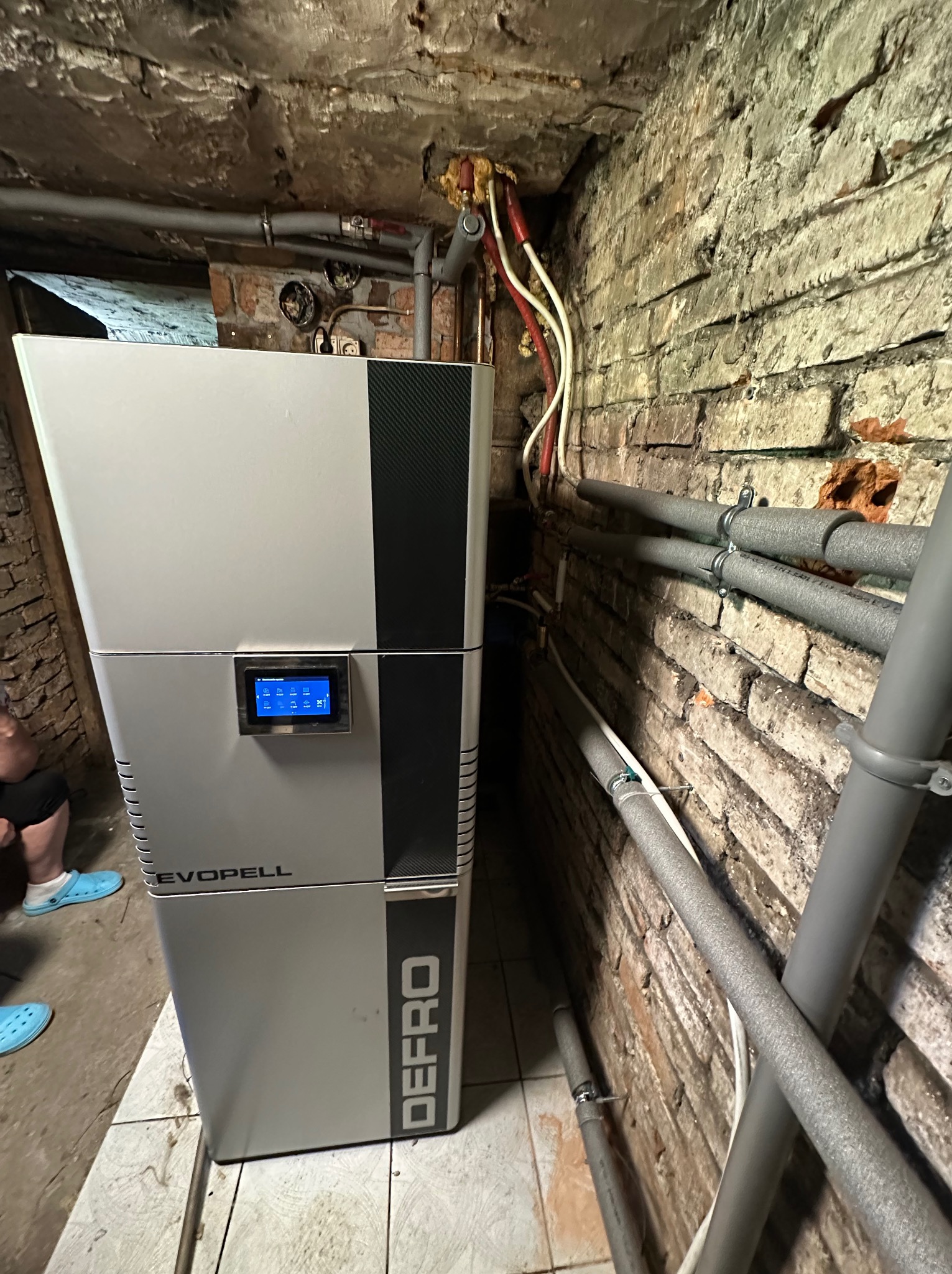 Nowoczesny piec DEFRO EVOPELL z dotykowym panelem sterowania, zainstalowany w piwnicy z odsłoniętą cegłą i rurami grzewczymi.