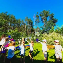 Grupa dzieci z pomponami i opiekunka na zielonym trawniku, w tle drzewa i błękitne niebo, sugerujące animacje na świeżym powietrzu.