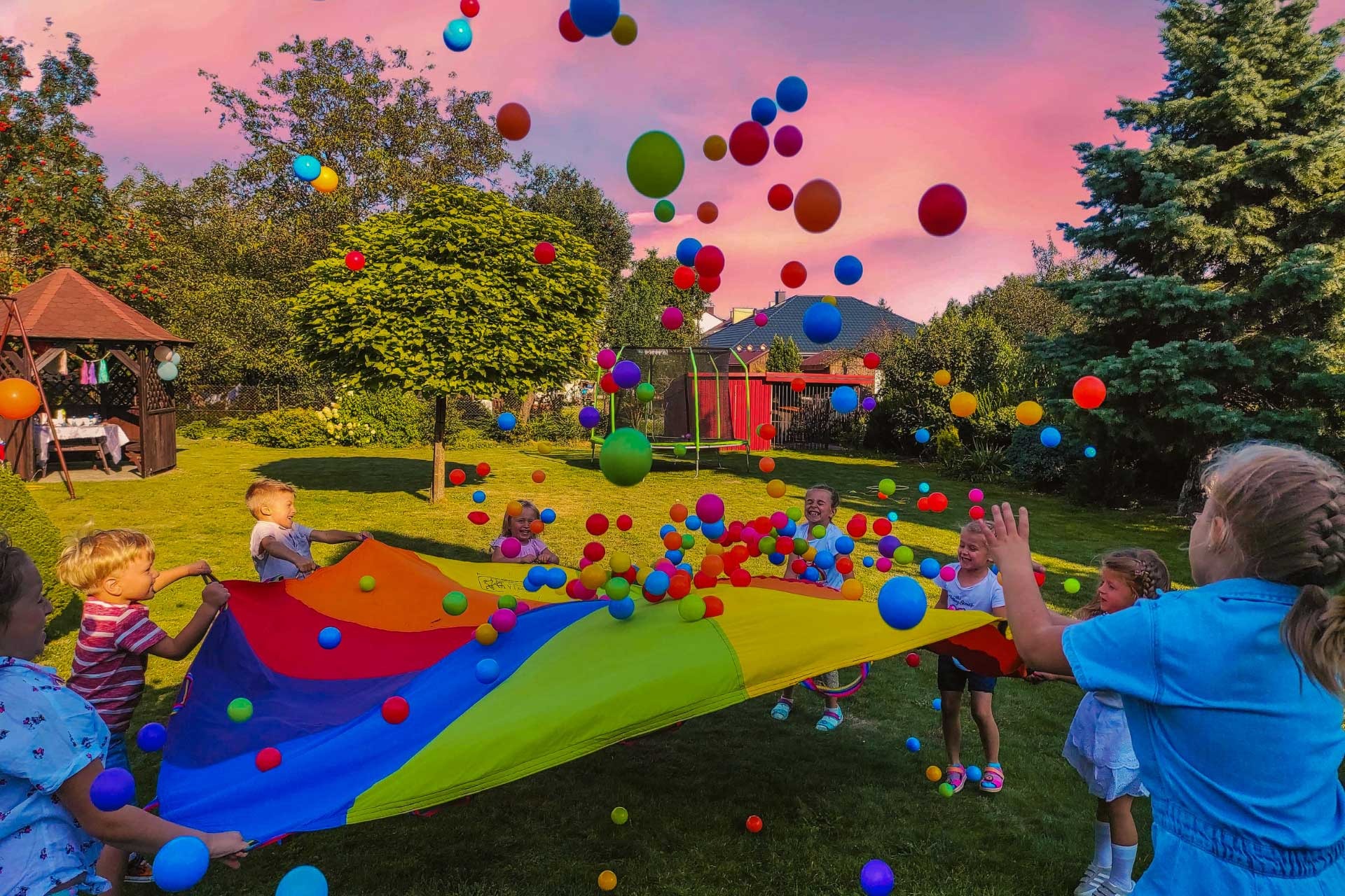 Grupa dzieci bawi się na trawie kolorową chustą animacyjną, podrzucając w górę mnóstwo kolorowych piłeczek, w tle widoczny ogród z altaną i trampoliną.