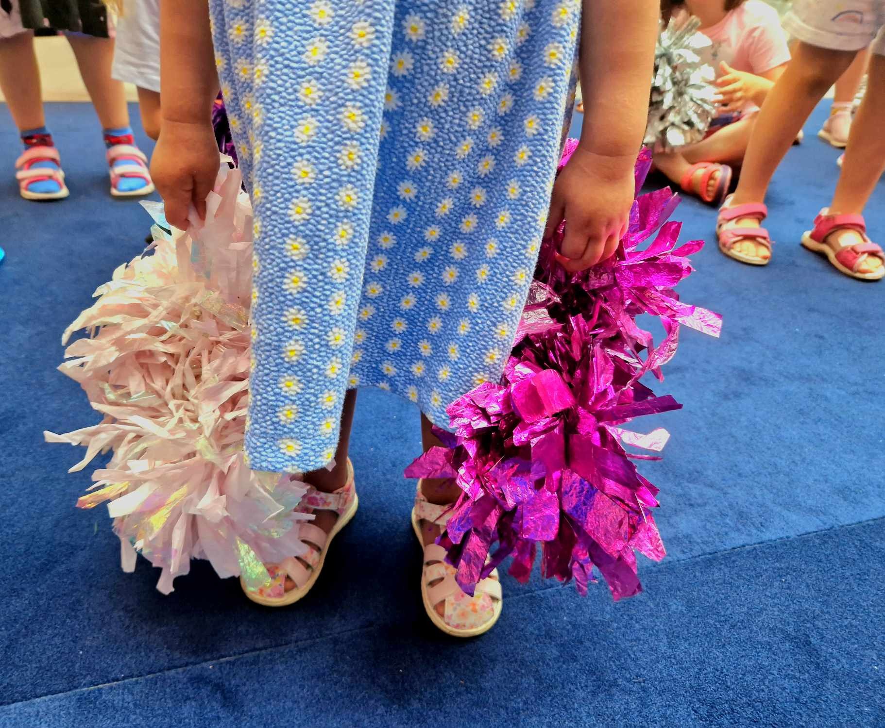 Dziecko w niebieskiej sukience w stokrotki trzyma dwa pompony: różowy i fioletowy, na niebieskiej podłodze, widoczne stopy innych dzieci.