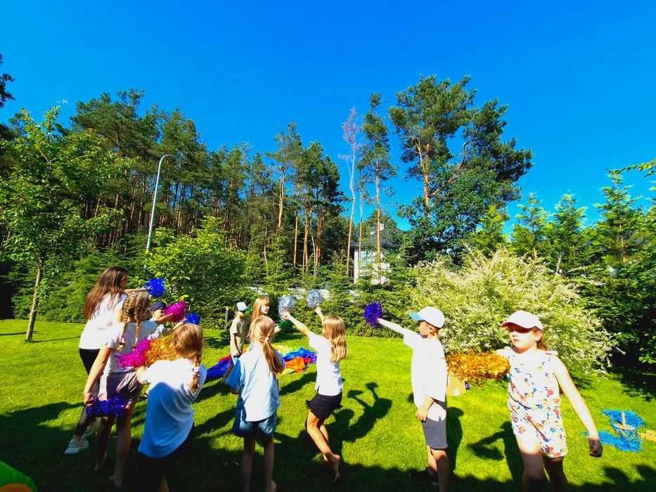 Grupa dzieci z pomponami i opiekunka na zielonym trawniku, w tle drzewa i błękitne niebo, sugerujące animacje na świeżym powietrzu.