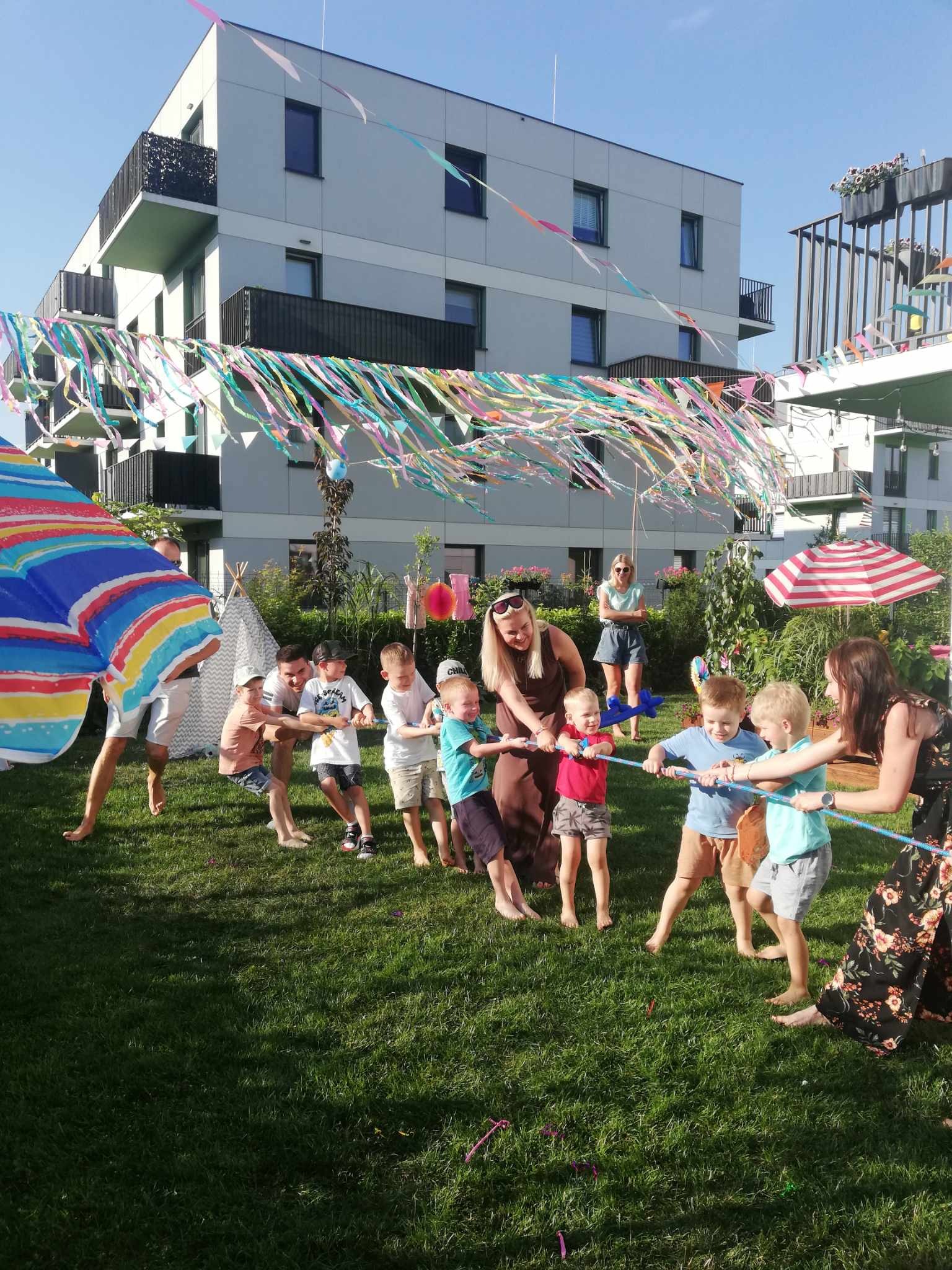 Grupa dzieci i dorosłych gra w przeciąganie liny na trawniku podczas imprezy plenerowej, w tle budynki mieszkalne i kolorowe dekoracje.