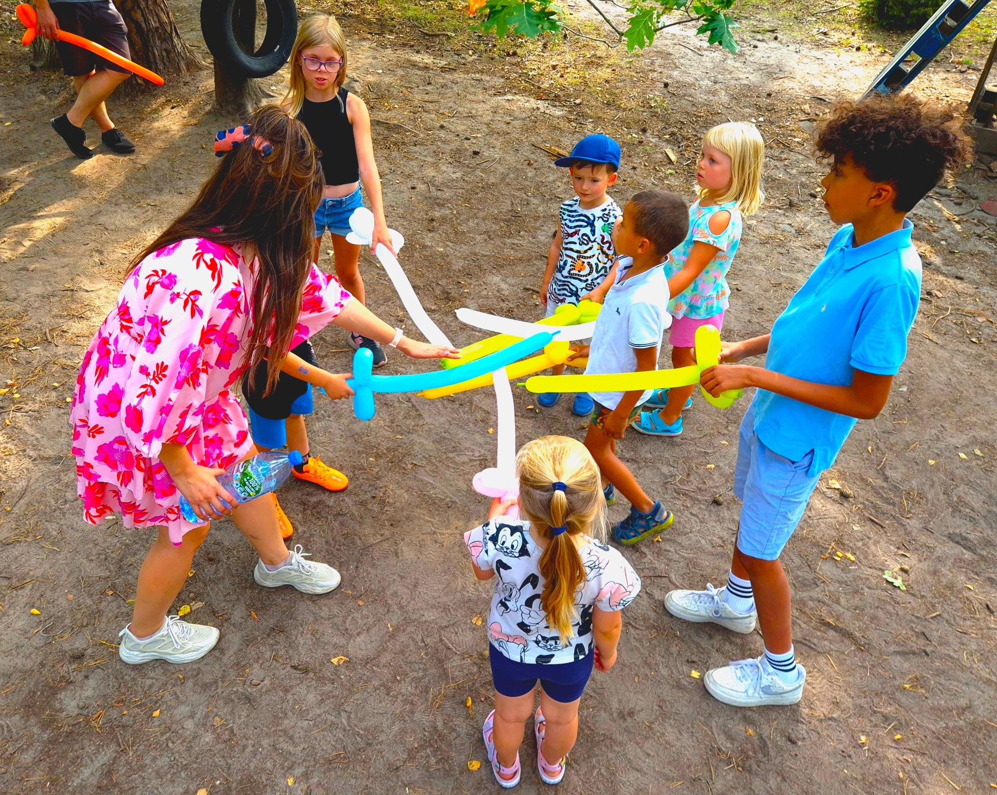 Grupa dzieci bawi się balonami modelowanymi na kształt mieczy pod okiem animatorki w kwiecistej sukience, w tle drzewa i opony.