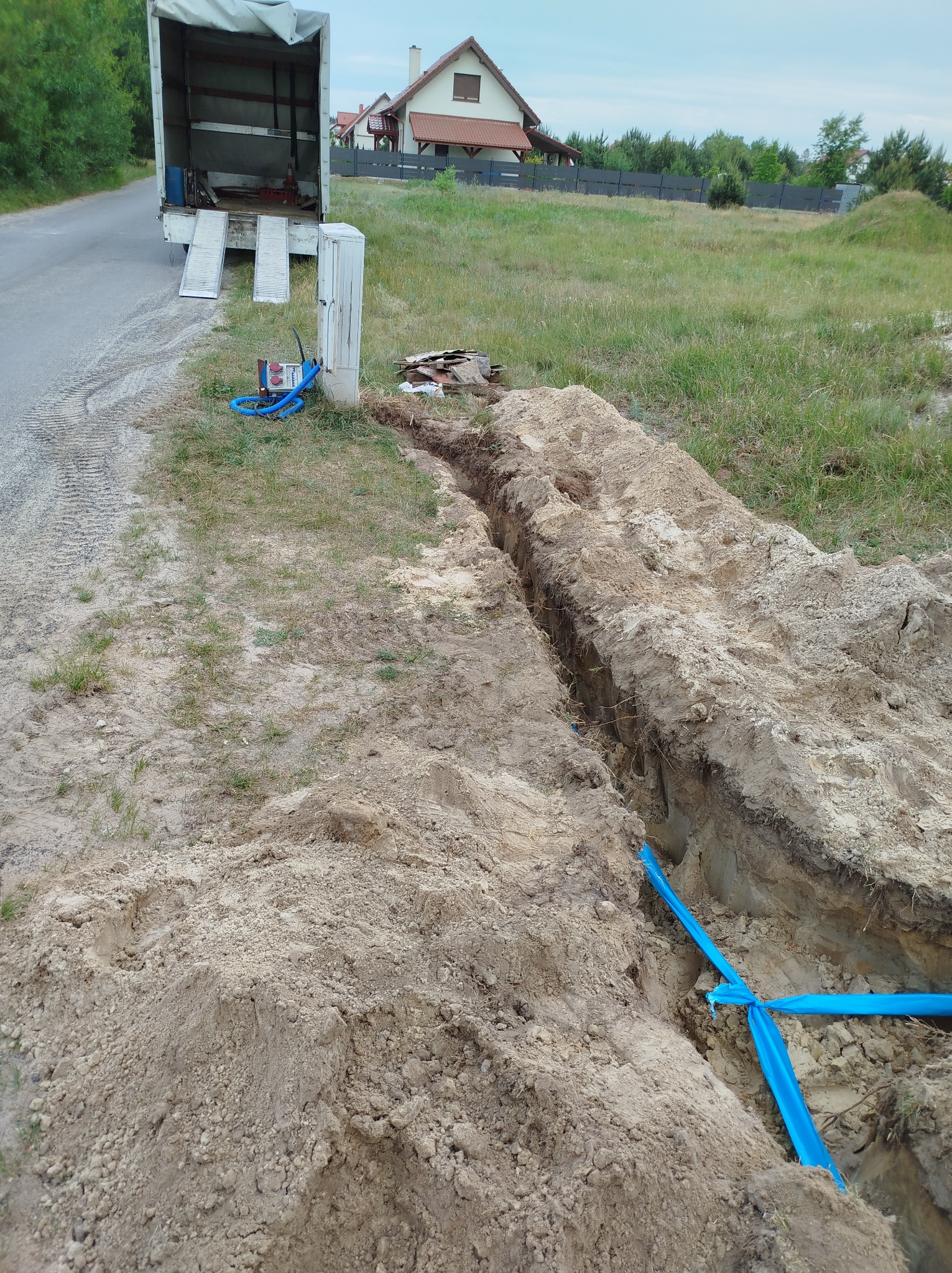 Wykop w ziemi z ułożonymi niebieskimi rurami, obok stoi skrzynka elektryczna i zaparkowany samochód dostawczy z otwartą rampą, w tle dom jednorodzinny.
