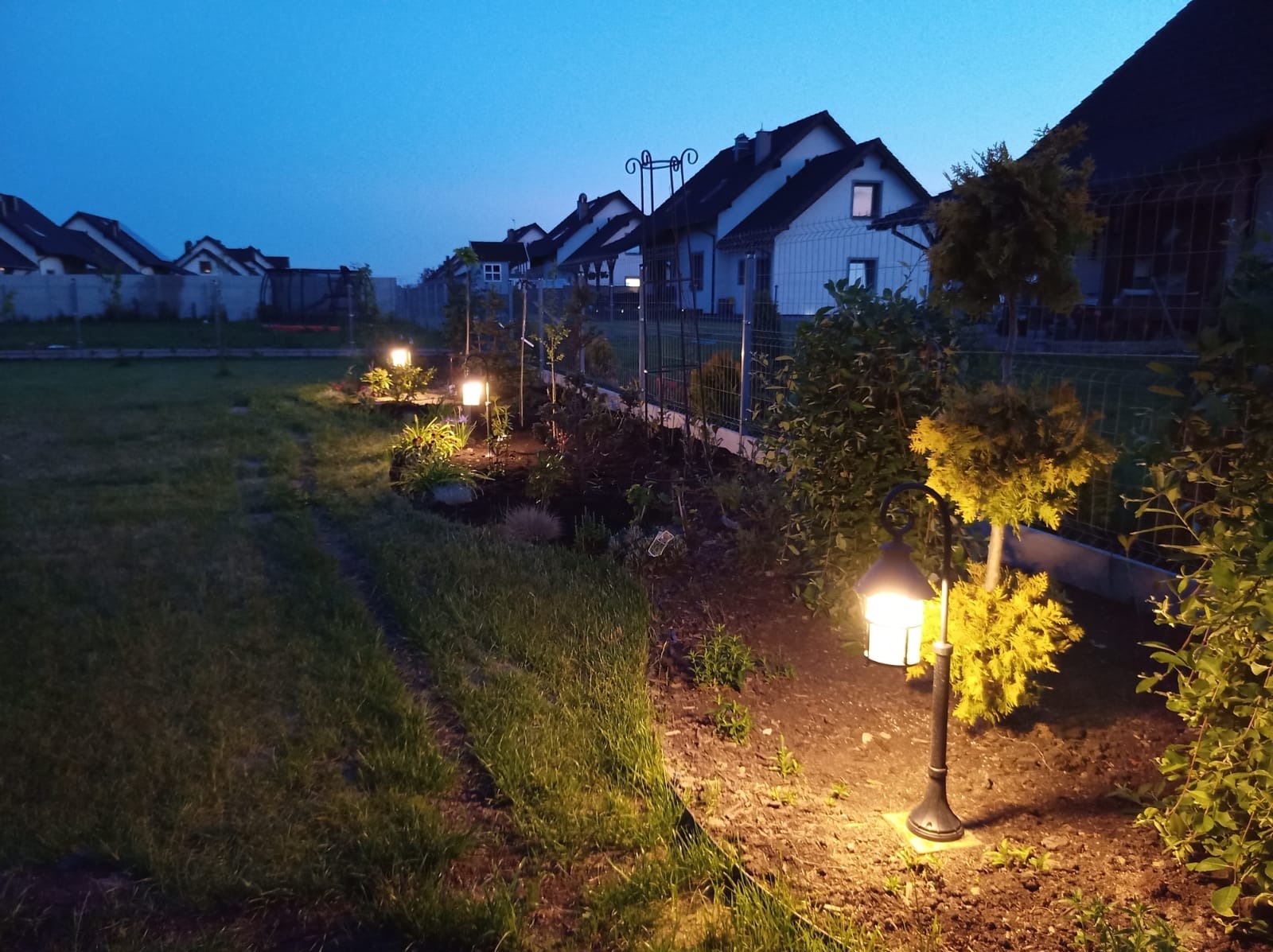 Oświetlony ogród wieczorową porą, widoczne lampy ogrodowe wzdłuż ścieżki i ogrodzenia, w tle domy jednorodzinne.