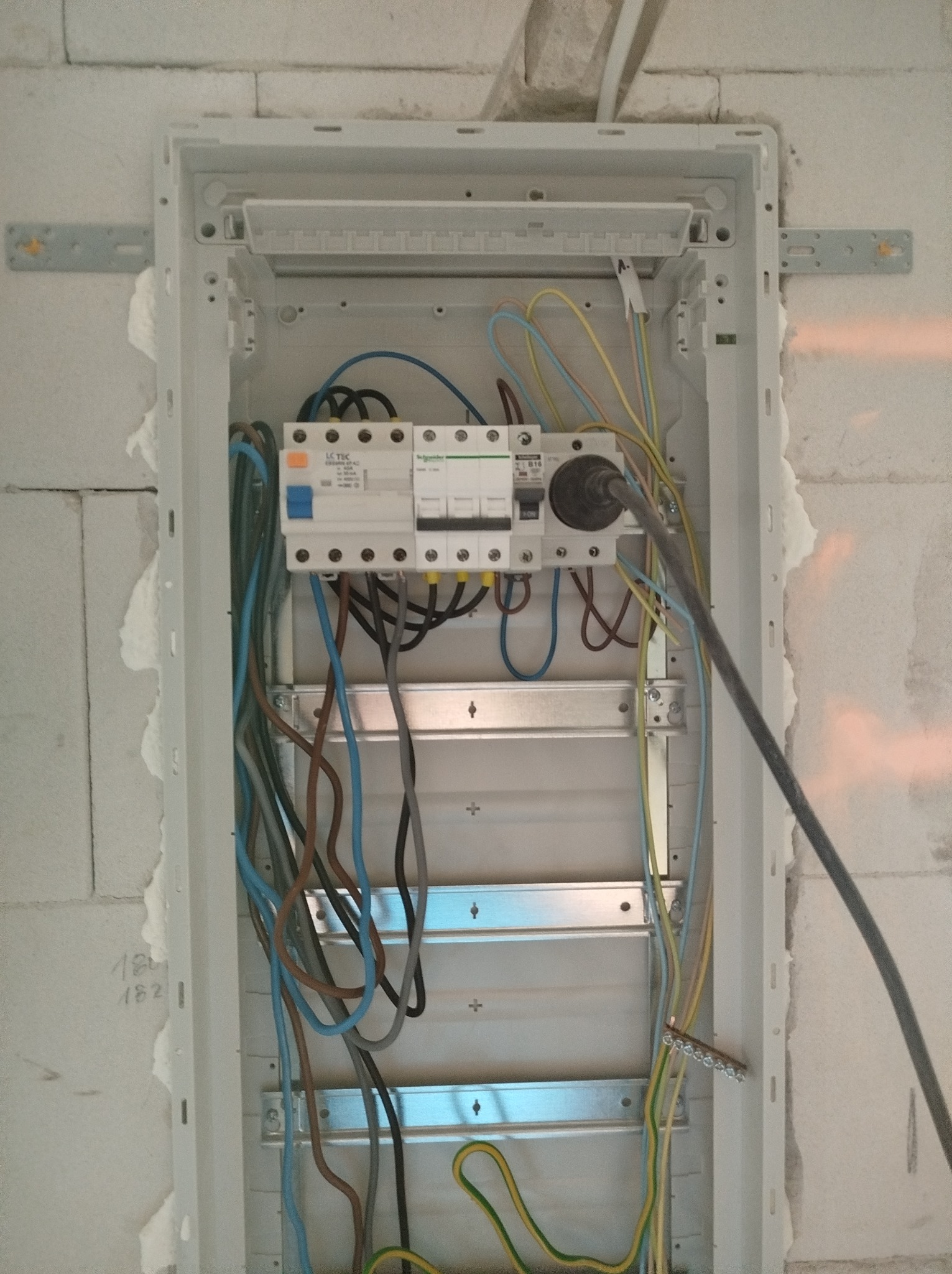 Szafa elektryczna w trakcie montażu, widoczne przewody, bezpieczniki, listwa zaciskowa i fragment ściany z bloku betonowego z zapisanymi numerami.