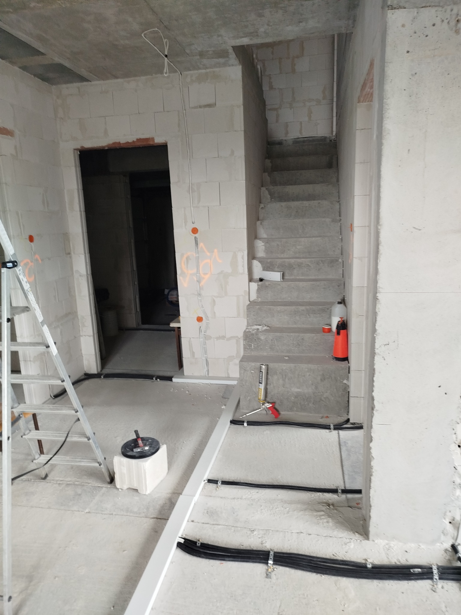 Surowy stan wykończenia wnętrza w trakcie prac instalacyjnych: widoczne kable elektryczne prowadzone wzdłuż ścian i podłogi, drabina aluminiowa, nieotynkowane ściany z betonu komórkowego...