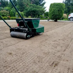 Zielony siewnik z wałem ugniatającym na świeżo obsianym gruncie, widoczne rzędy nasion w ziemi, w tle drzewa i krzewy.