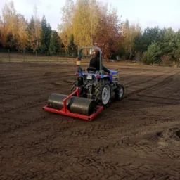 Niebieski traktor z walcem ugniata ziemię na dużej powierzchni pola, widoczne ślady opon, w tle drzewa z jesiennymi liśćmi.
