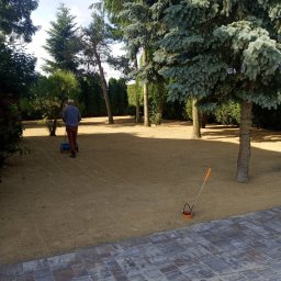 GARDEN OF SUCESS Magdalena Tołysz - Człowiek z siewnikiem na piaskowym podłożu, przygotowanym pod trawnik. W tle drzewa iglaste i liściaste. W rogu widoczny fragment kostki brukowej.