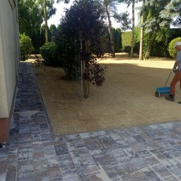 GARDEN OF SUCESS Magdalena Tołysz - Przygotowany teren pod trawnik z rolki, widoczny fragment ułożonej kostki brukowej i osoba z wózkiem do siewu na tle zieleni ogrodowej w słoneczny dzień.