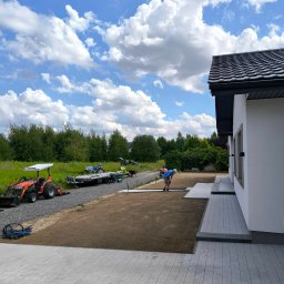 GARDEN OF SUCESS Magdalena Tołysz - Układanie kostki brukowej na posesji domu jednorodzinnego. Widoczny traktor, przygotowany teren i pracownik w trakcie pracy. Błękitne niebo z chmurami w tle.