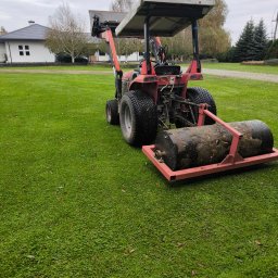 GARDEN OF SUCESS Magdalena Tołysz - Czerwony traktor z wałem ugniatającym na zielonym trawniku, w tle dom z szarą dachówką i drzewa. Widok z tyłu na maszynę do pielęgnacji trawnika.