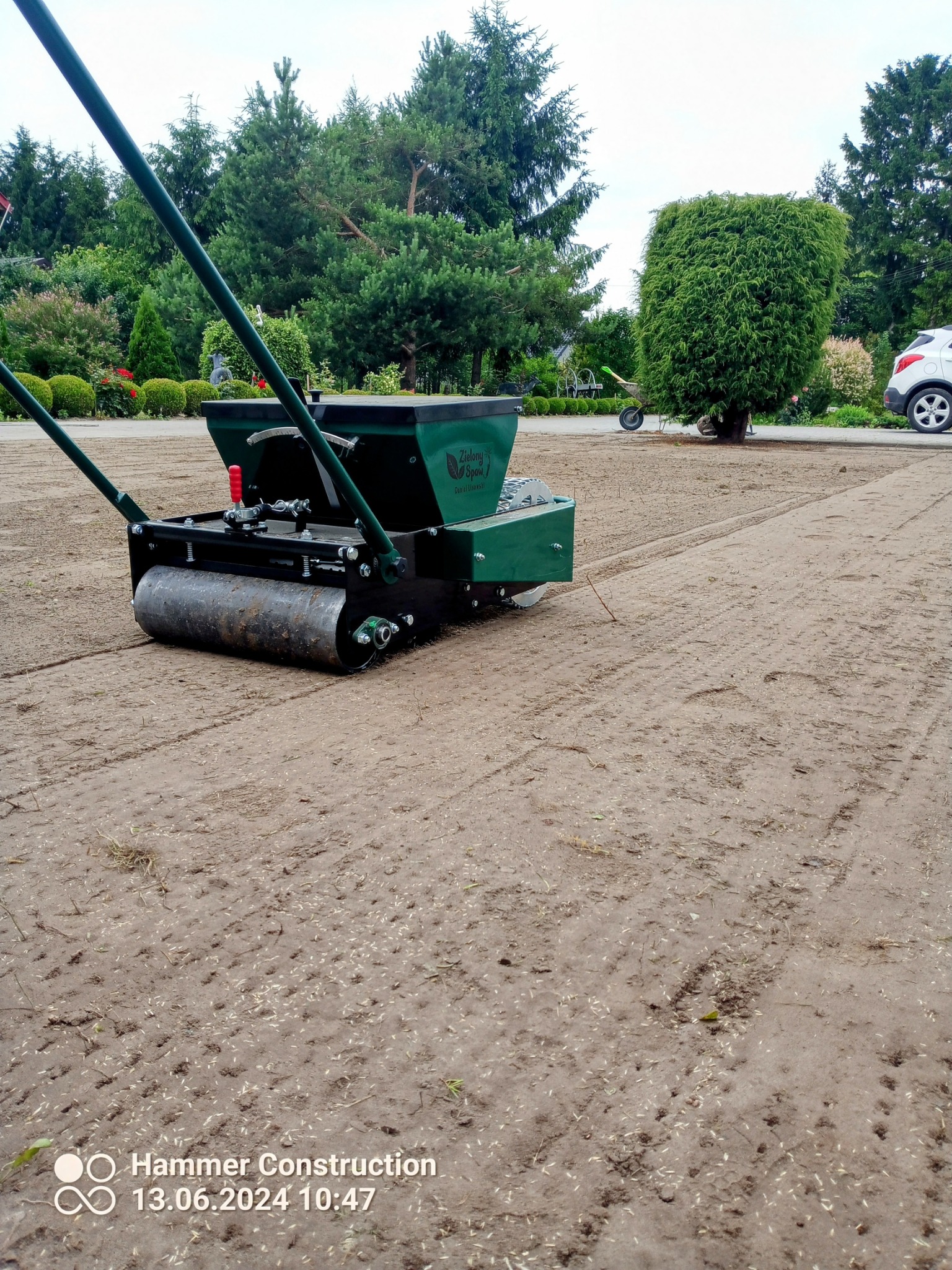 Zielony siewnik z wałem ugniatającym na świeżo obsianym gruncie, widoczne rzędy nasion w ziemi, w tle drzewa i krzewy.