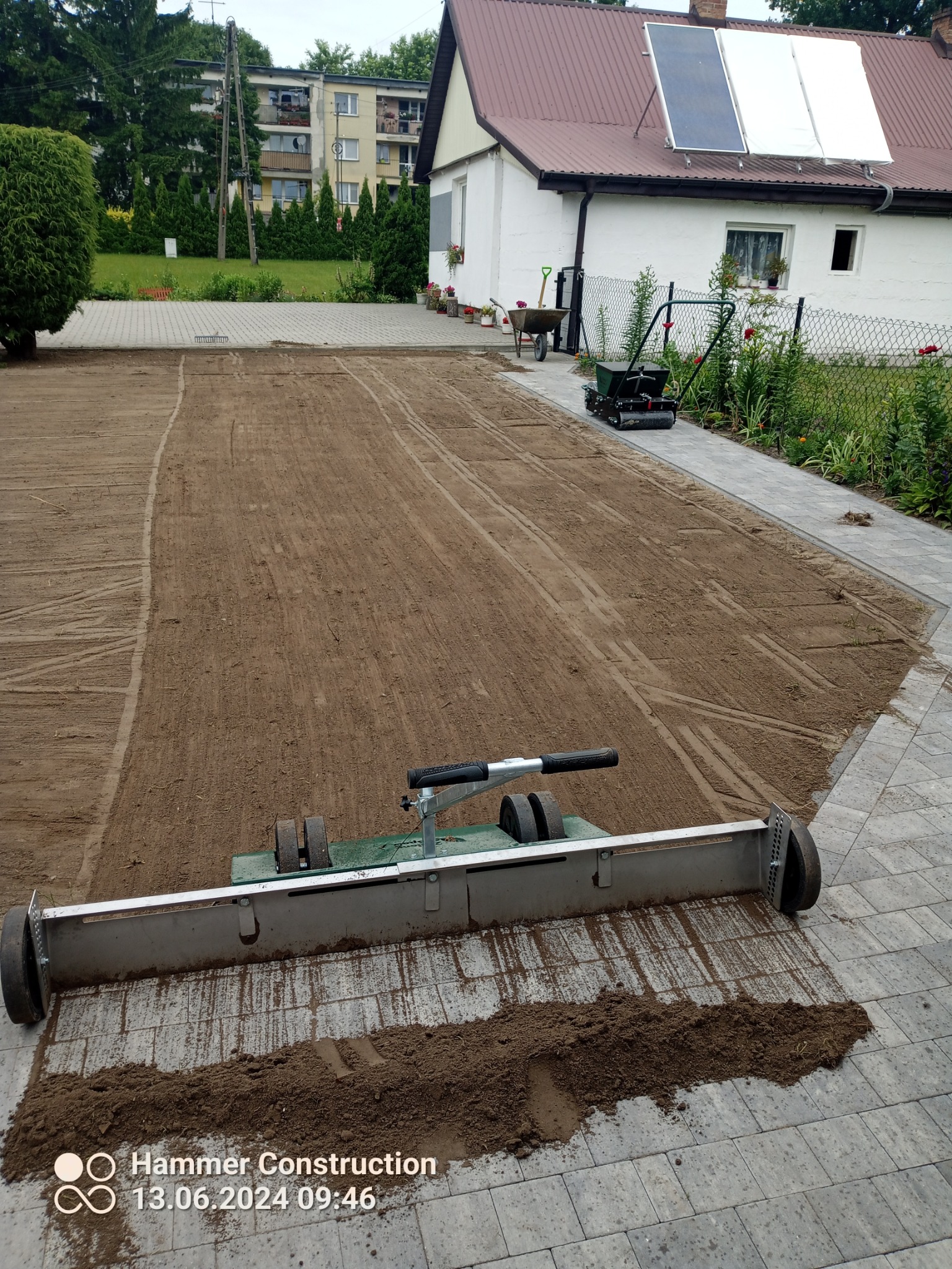 Przygotowanie terenu pod trawnik: wyrównana powierzchnia ziemi z widocznymi śladami po maszynie do wyrównywania, w tle dom z panelami solarnymi i narzędzia ogrodnicze.