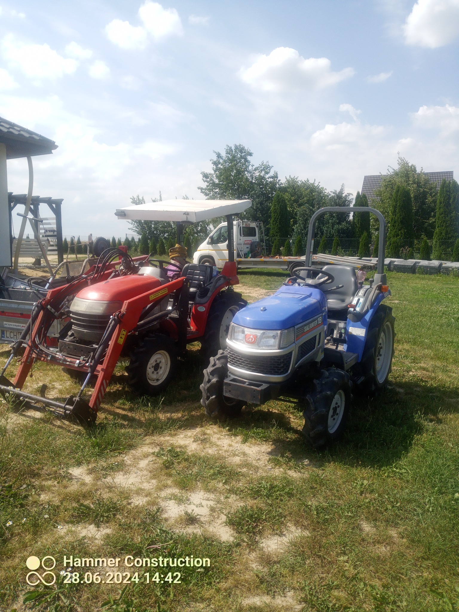 Dwa małe traktory, czerwony z ładowaczem czołowym i niebieski, stoją na trawiastym terenie przed budynkiem i samochodem dostawczym w słoneczny dzień.