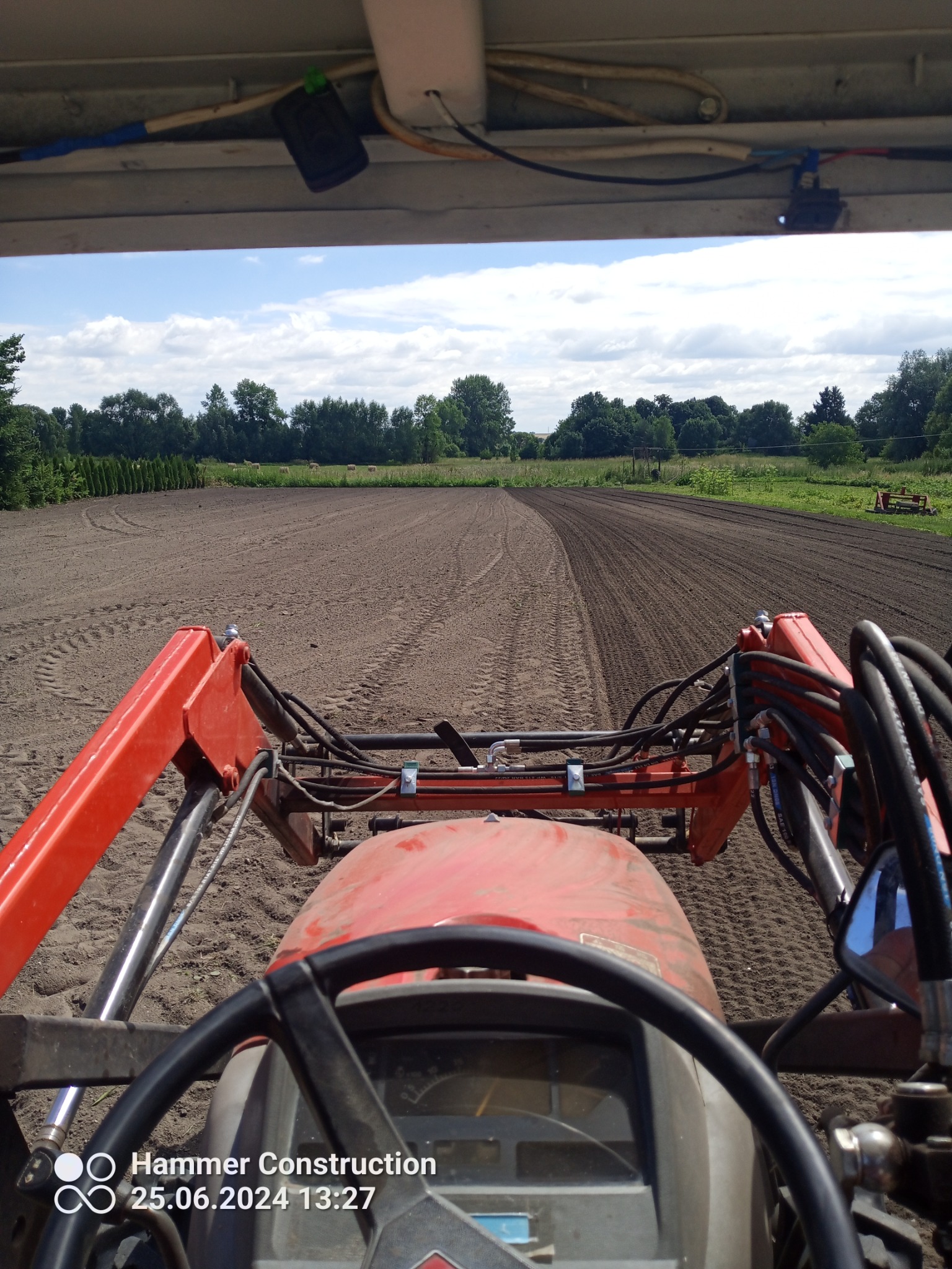 Widok z kabiny traktora na świeżo wyrównany teren pod przyszły ogród, z widocznymi śladami opon i redlic, drzewa w tle.