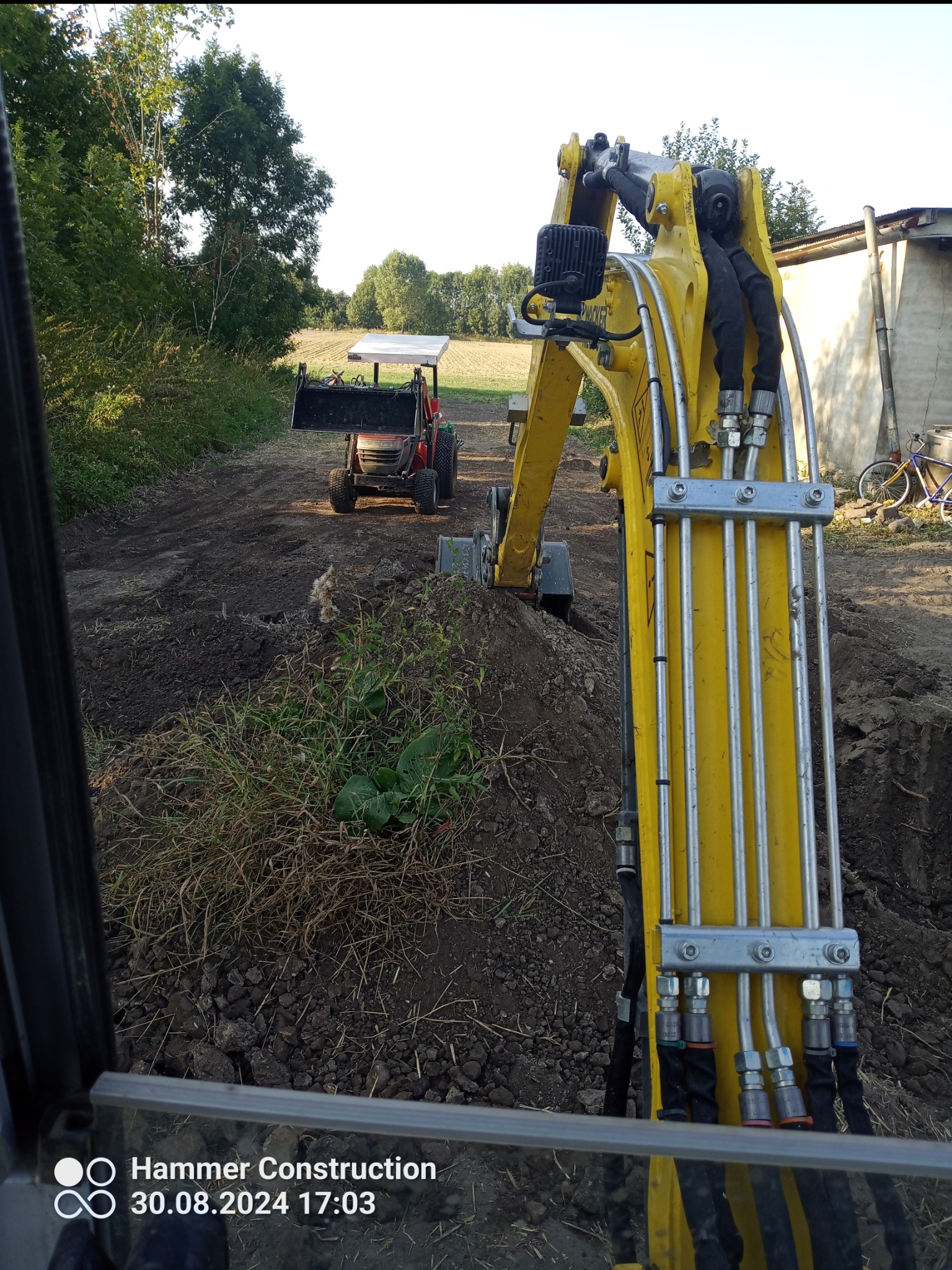 Widok z kabiny na żółtą mini-koparkę Wacker Neuson podczas prac ziemnych, w tle widoczny mały traktor z ładowaczem czołowym, teren częściowo wyrównany.
