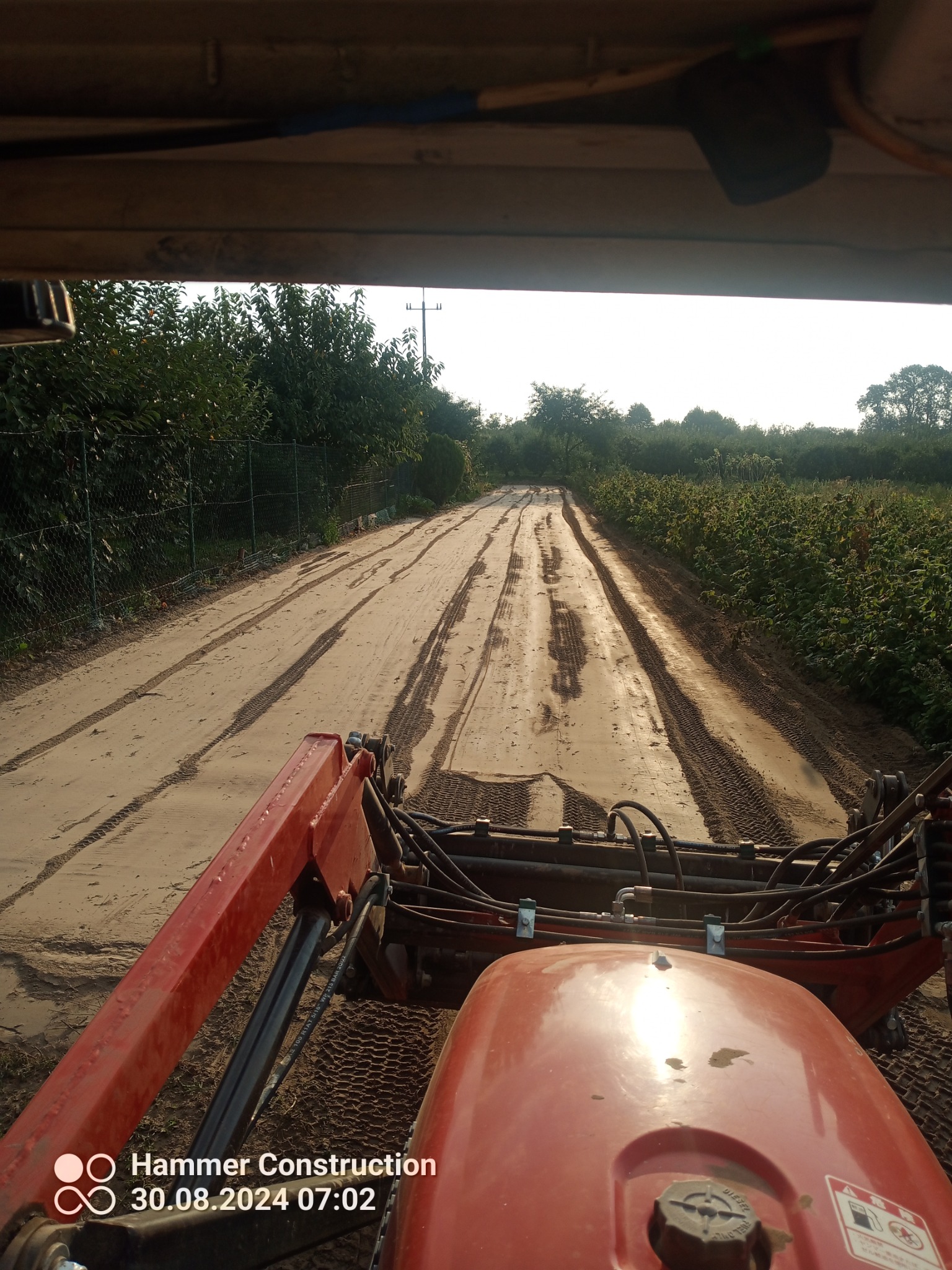 Widok z kabiny traktora na wyrównaną, piaszczystą drogę gruntową pośród zieleni, ślady opon widoczne w piasku, w oddali słup energetyczny.