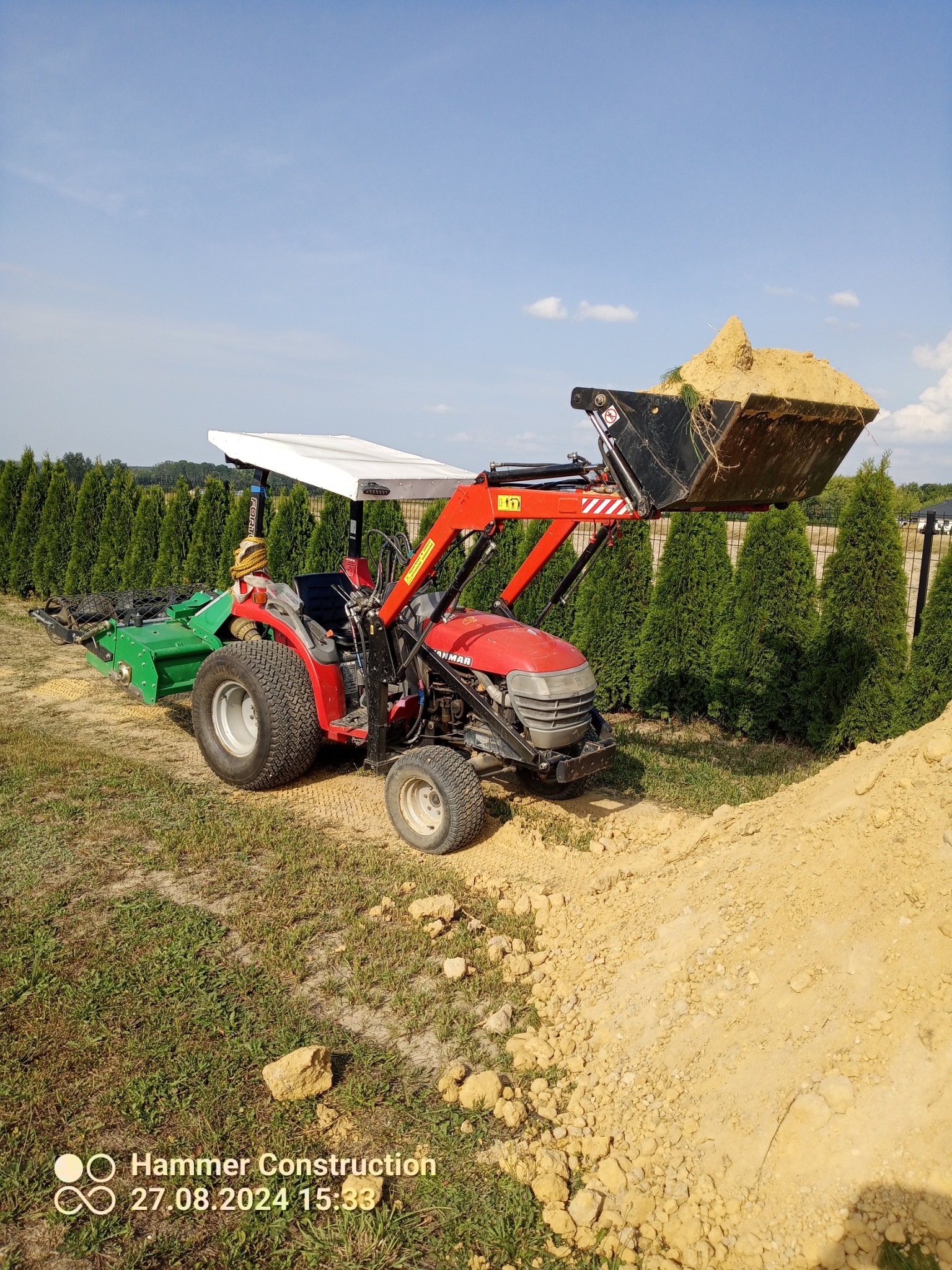 Czerwony traktor Yanmar z ładowarką pełną piasku i glebogryzarką, na tle rzędu tuj w ogrodzie.