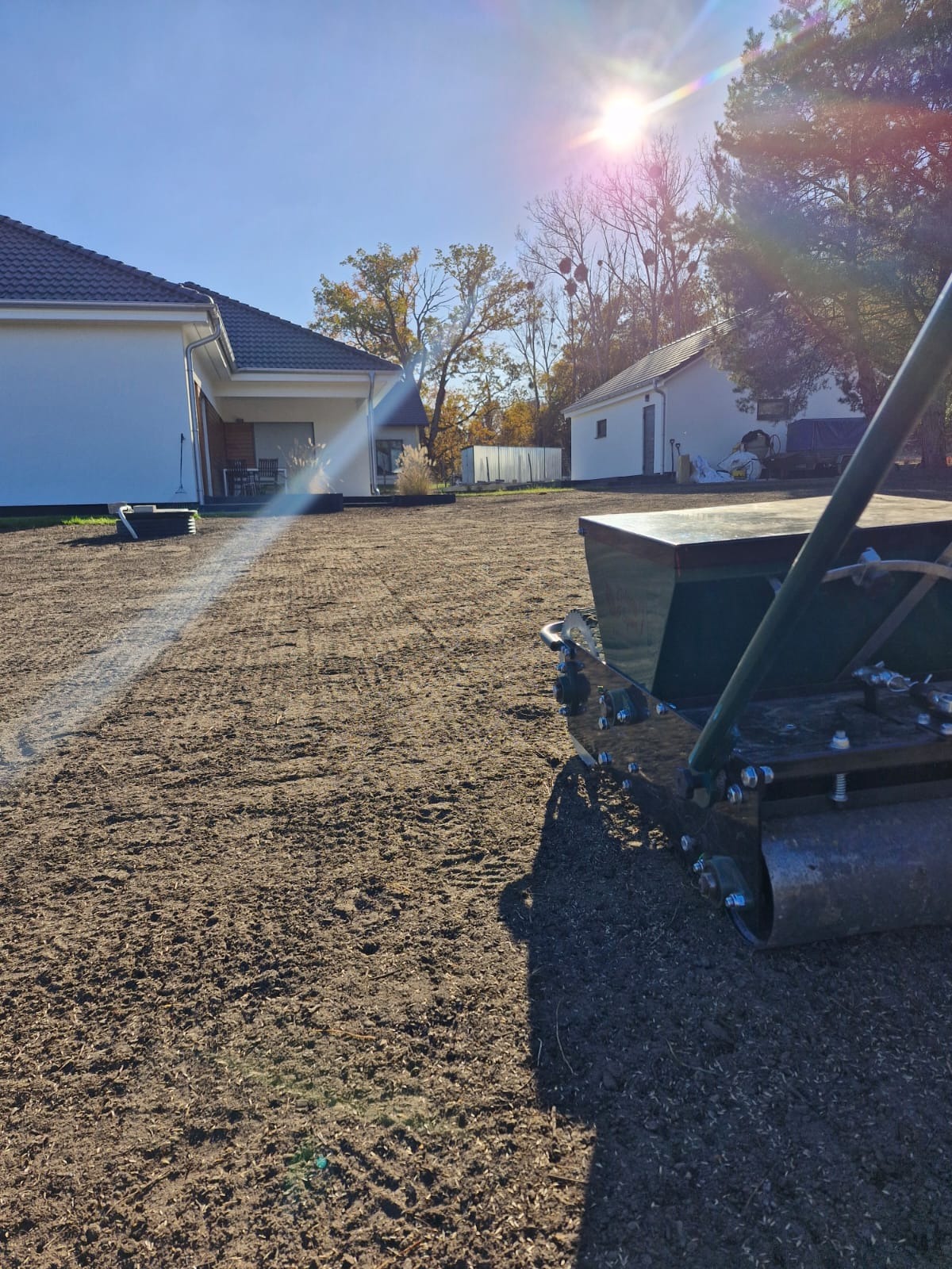 Niwelacja terenu pod trawnik siewnikiem, widoczne ślady po przejeździe, domy w tle, słoneczny dzień.