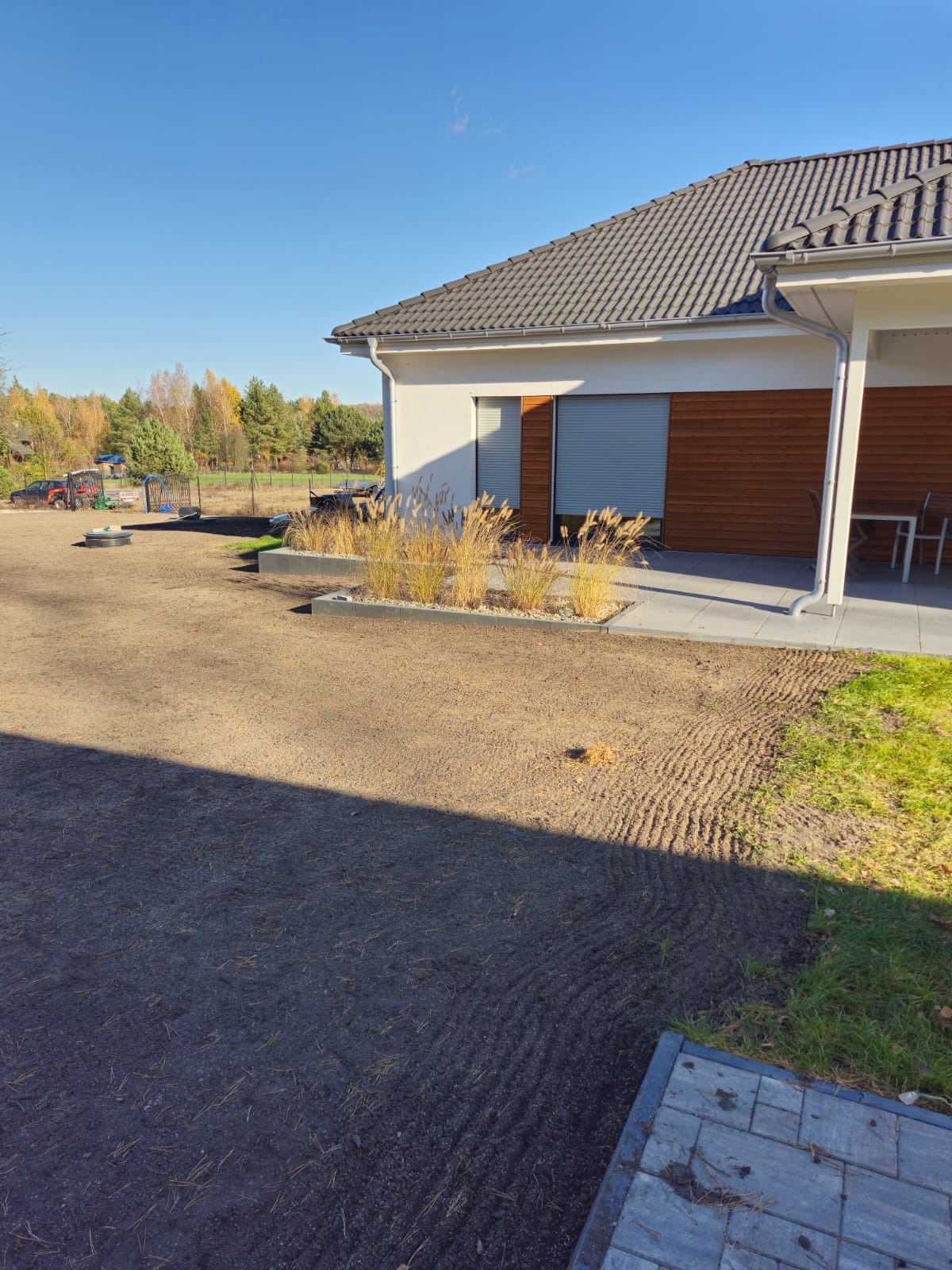 Przygotowany teren pod trawnik z widocznymi śladami po bronowaniu, obok rabata z ozdobnymi trawami i fragment domu z drewnianą elewacją.