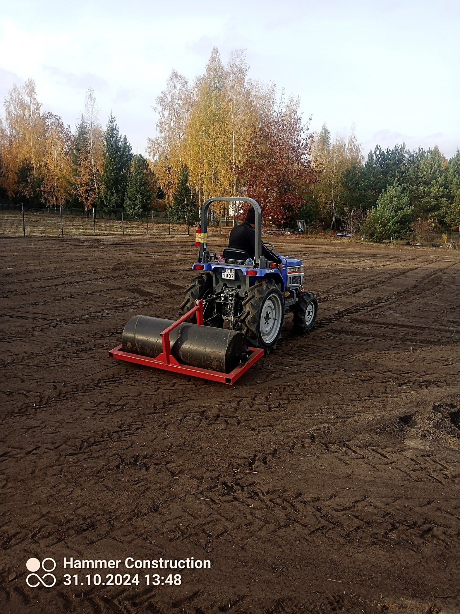 Niebieski traktor z walcem ugniata ziemię na dużej powierzchni pola, widoczne ślady opon, w tle drzewa z jesiennymi liśćmi.