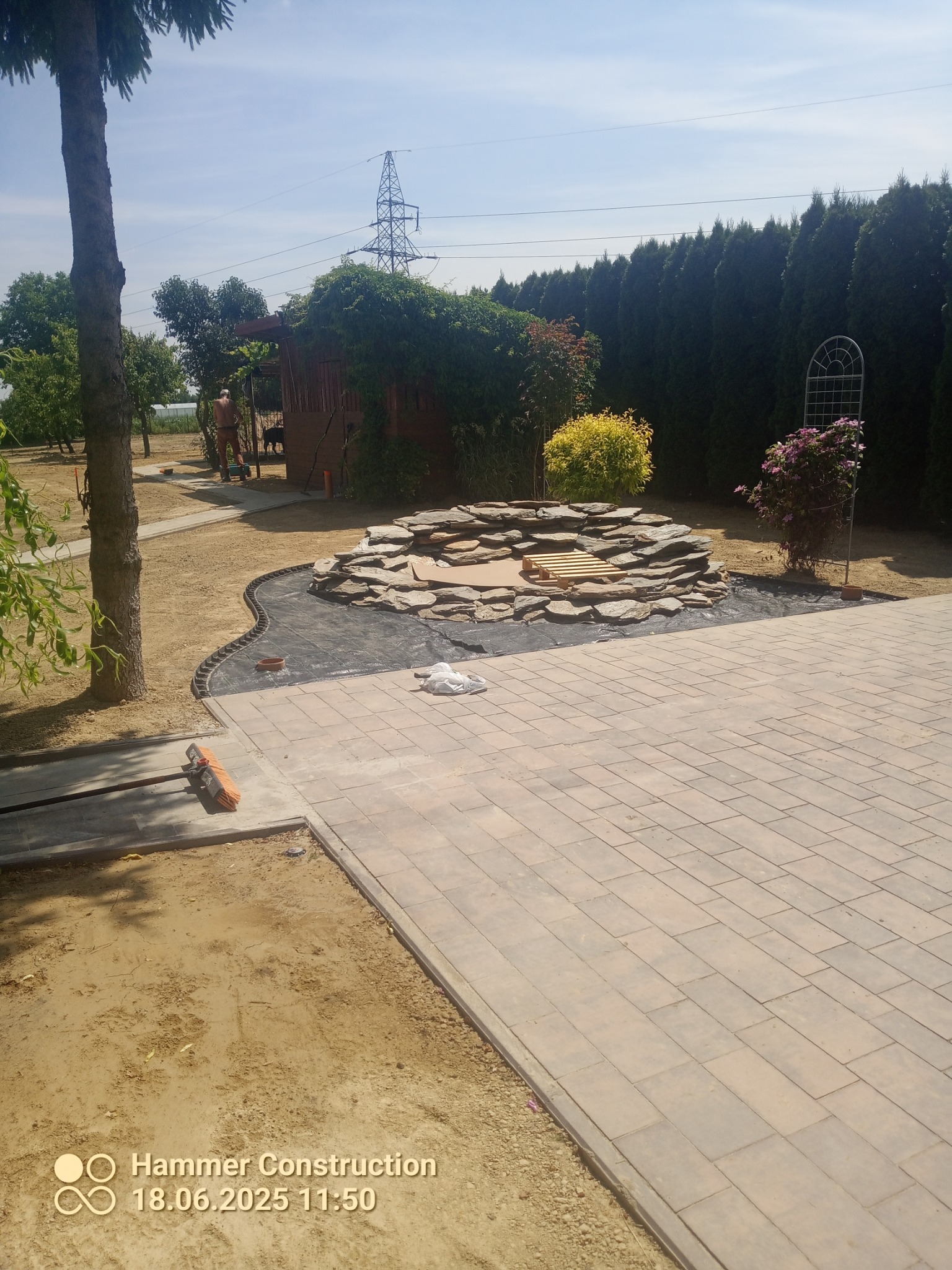 Układanie kostki brukowej na posesji z oczkiem wodnym w tle. Widoczny fragment ogrodu, drzewa i altana. Bruk w odcieniach szarości, regularny wzór.