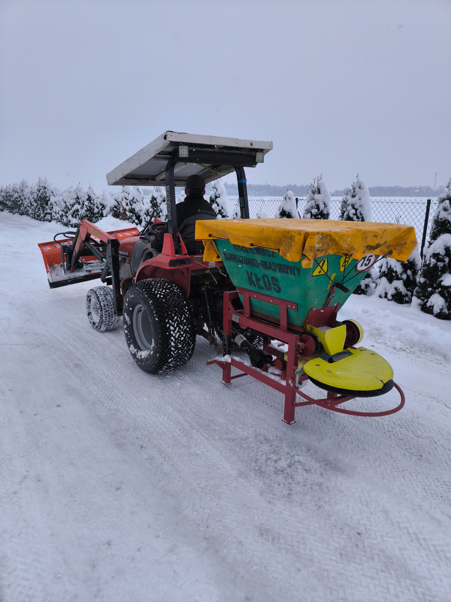 Czerwony traktor z rozsiewaczem Kłos na zaśnieżonej drodze, otoczony ośnieżonymi drzewami iglastymi. Widoczny operator w kabinie. Zima.