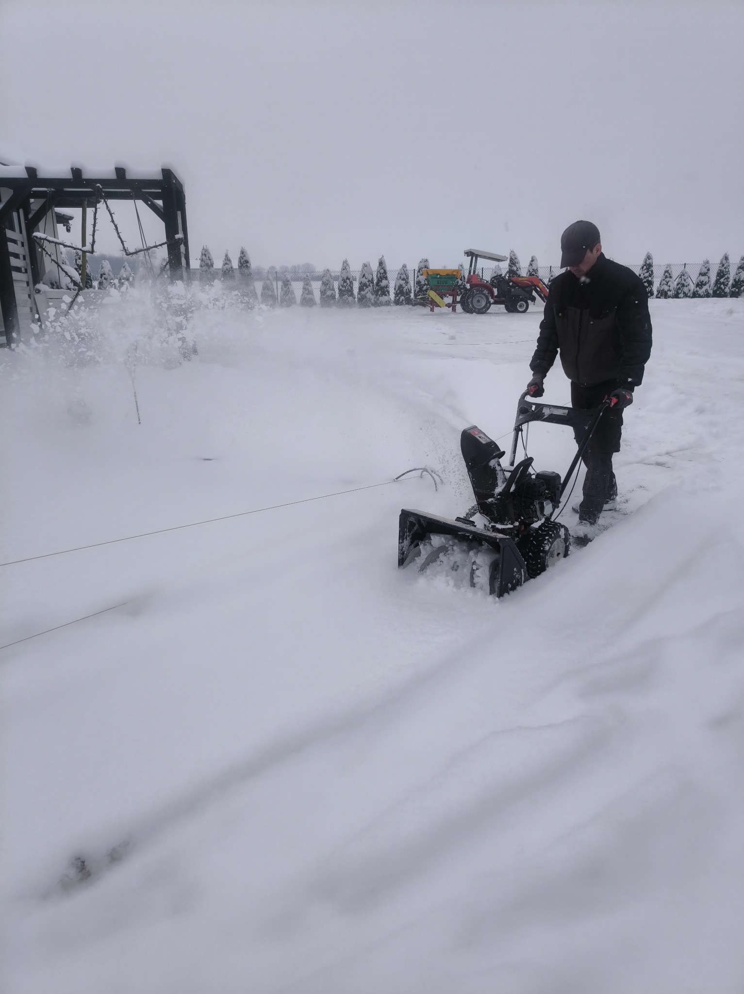 Mężczyzna obsługuje odśnieżarkę spalinową na zasypanym śniegiem terenie. W tle widoczny traktor i rząd iglaków.