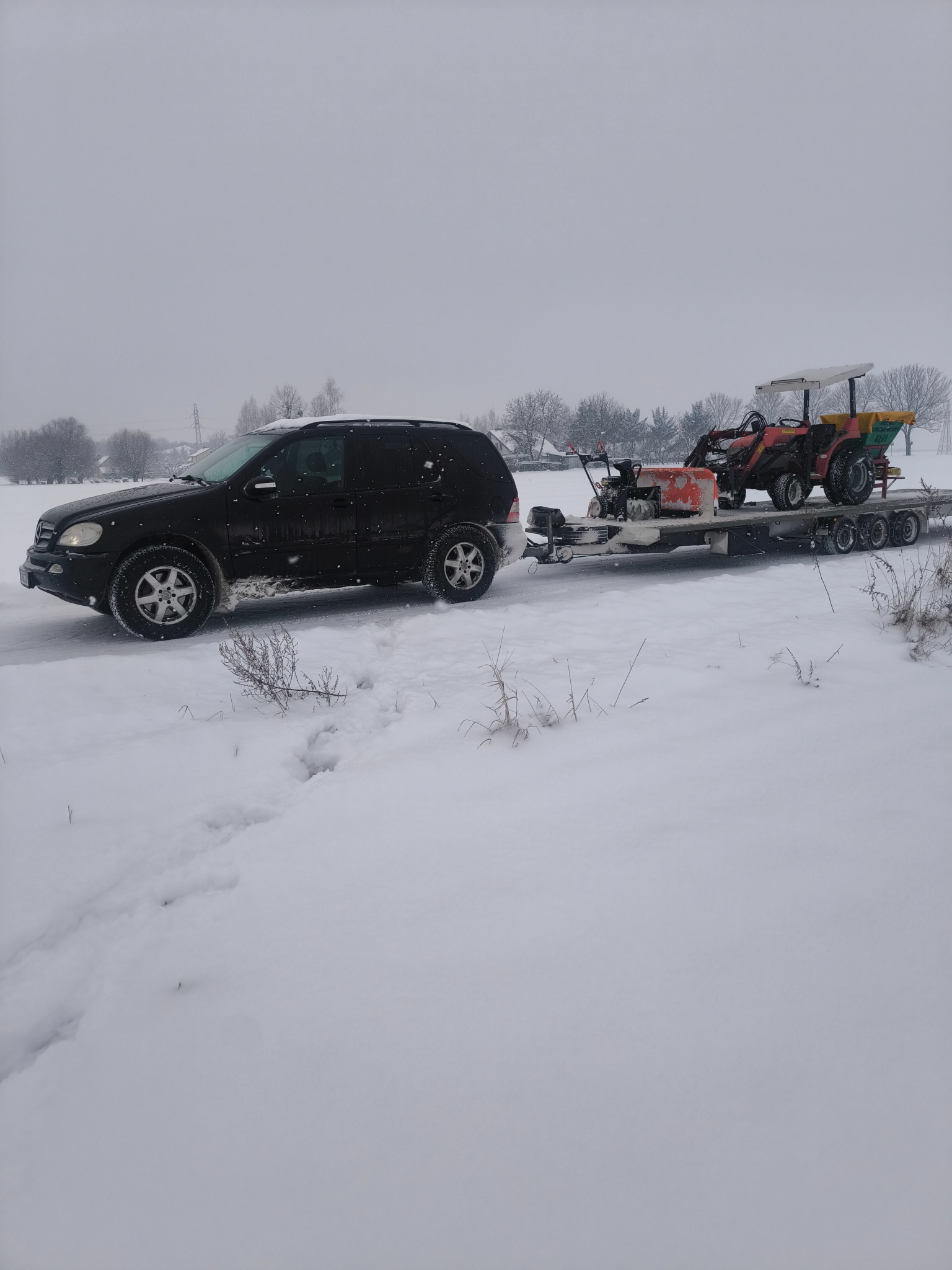 Czarny SUV ciągnie przyczepę z traktorem i odśnieżarką na zaśnieżonym terenie. Zimowy krajobraz, szare niebo.