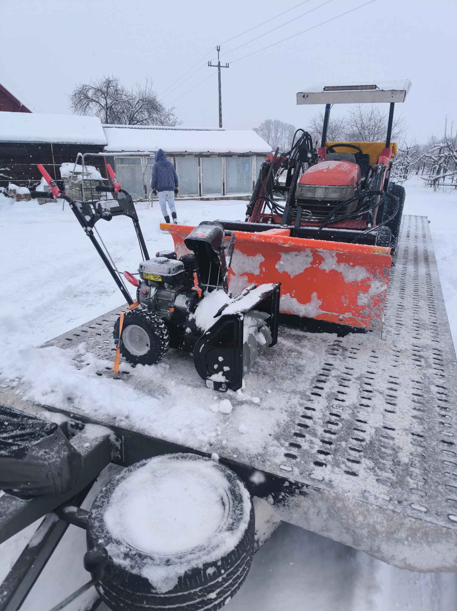 Dwa odśnieżarki, w tym traktor z pługiem, stoją na platformie przyczepy w zimowej scenerii. W tle budynki i osoba w kurtce.