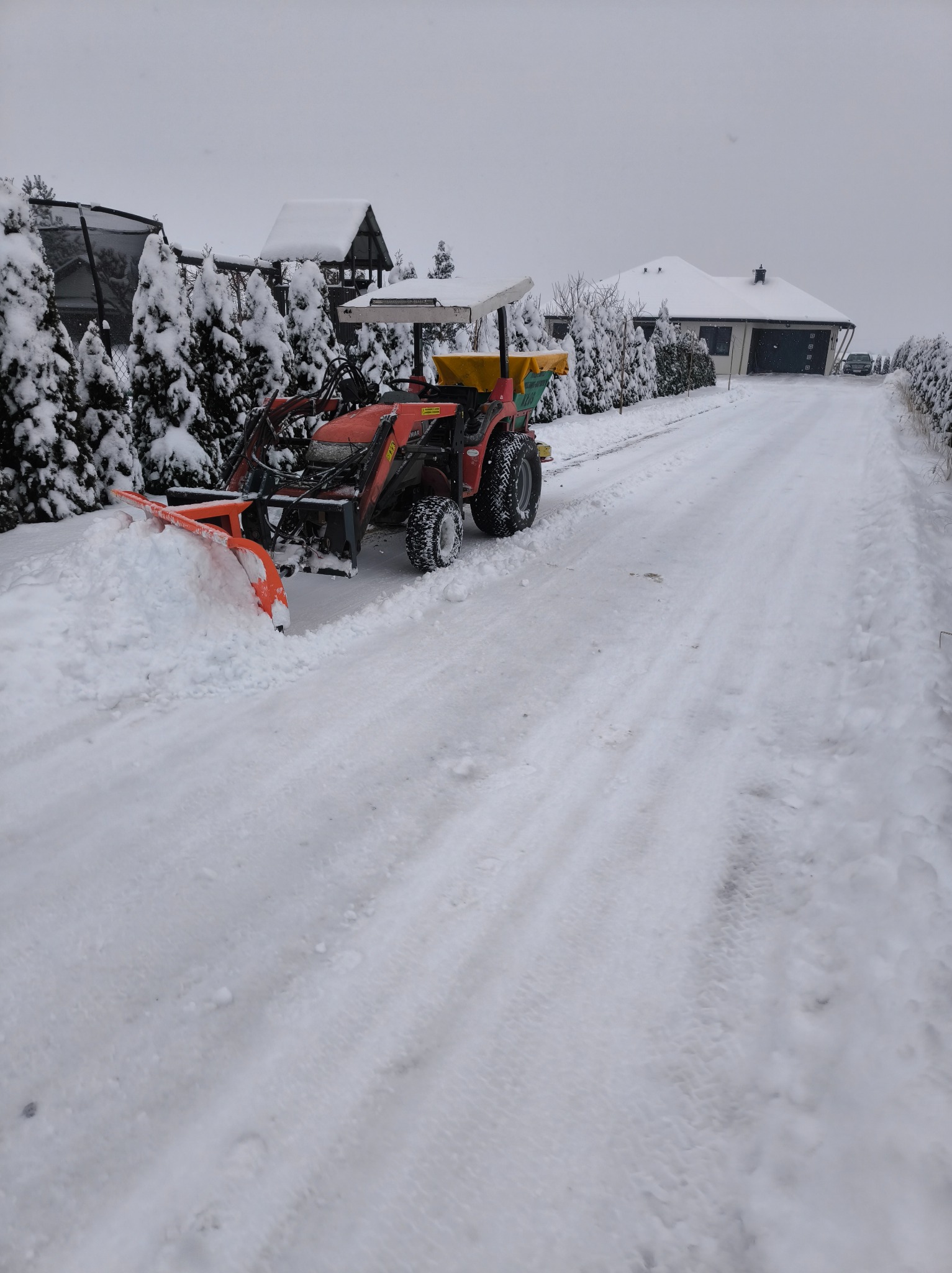 Czerwony traktor z pługiem odśnieża zasypaną śniegiem drogę w Wierzbicy. Śnieg leży na trawnikach i dachach domów.