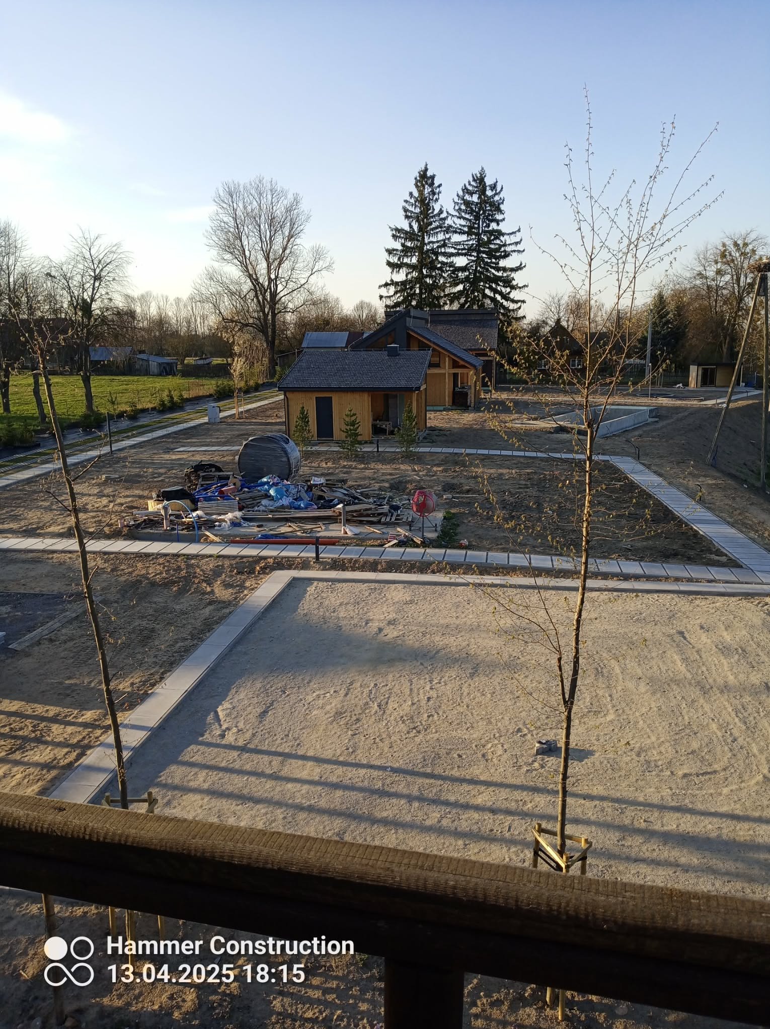 Plac budowy z elementami ogrodu: ścieżki z kostki, piasek, młode drzewka. W tle drewniane budynki i drzewa iglaste. Widok z góry, wczesna faza prac.