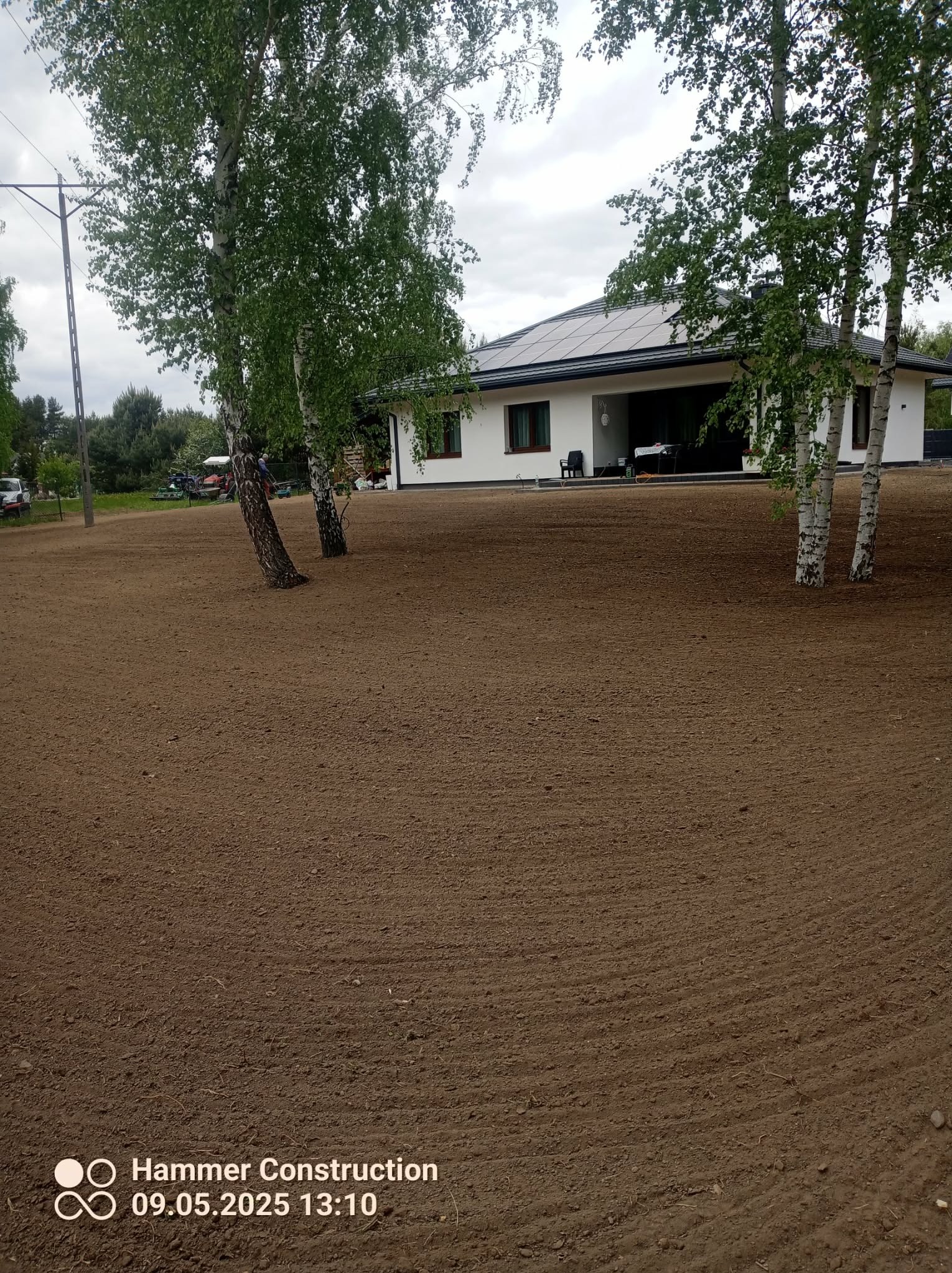 Przygotowany teren pod trawnik z widocznymi śladami po bronowaniu, kilka brzóz na działce, w tle dom z panelami słonecznymi. Dzień pochmurny.