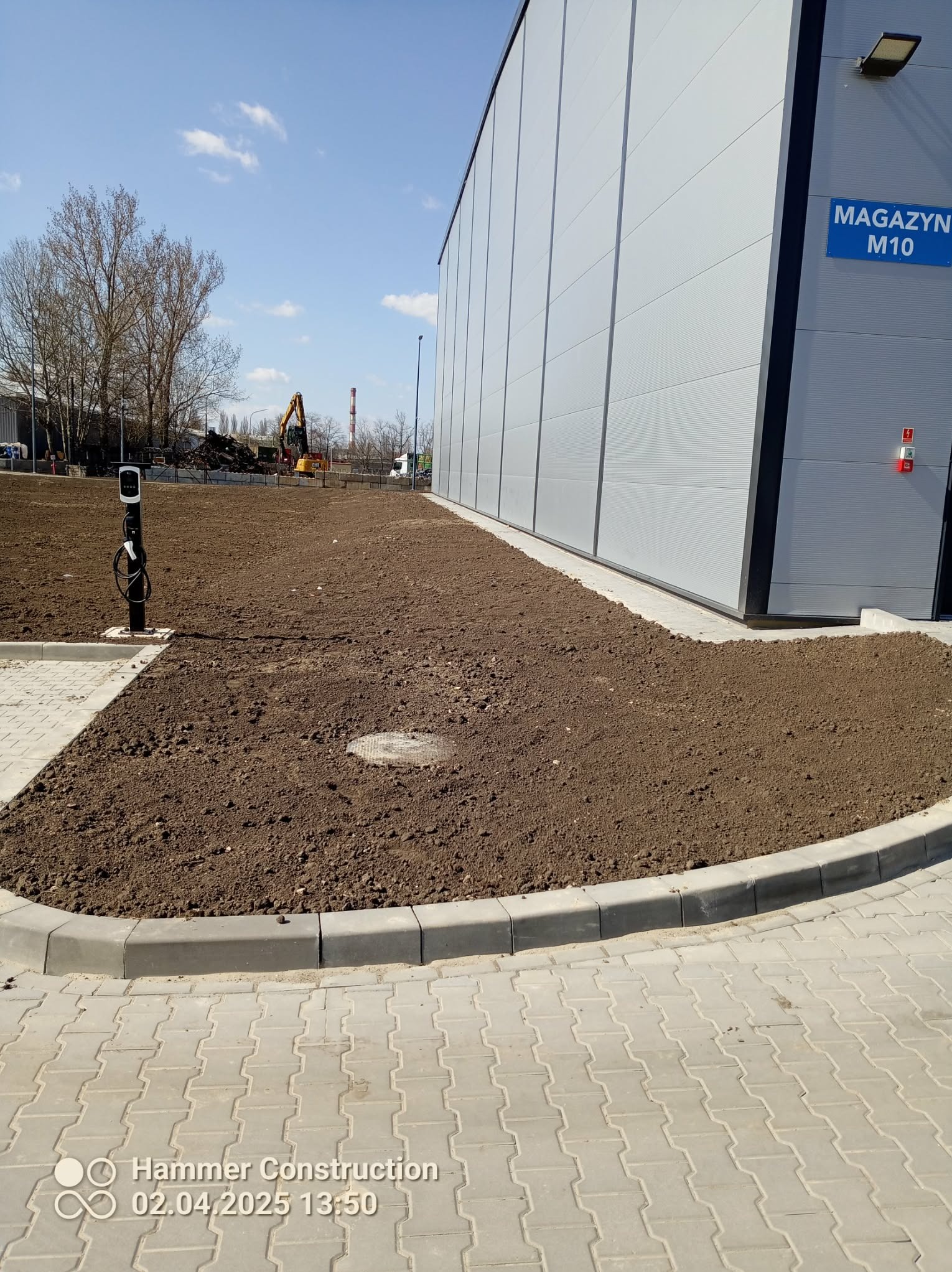 Przygotowany teren pod trawnik przy budynku magazynowym z kostką brukową i obrzeżami. W tle koparka i hałda ziemi.