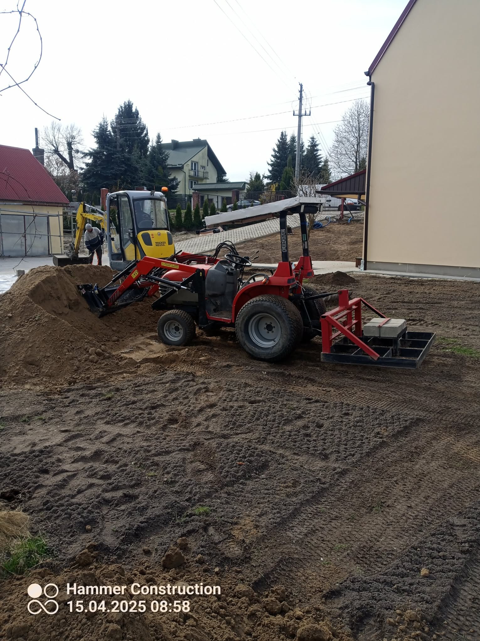 Czerwony traktor z osprzętem i żółta koparka na placu budowy. W tle domy jednorodzinne i drzewa. Prace ziemne w toku, przygotowanie podłoża pod budowę.