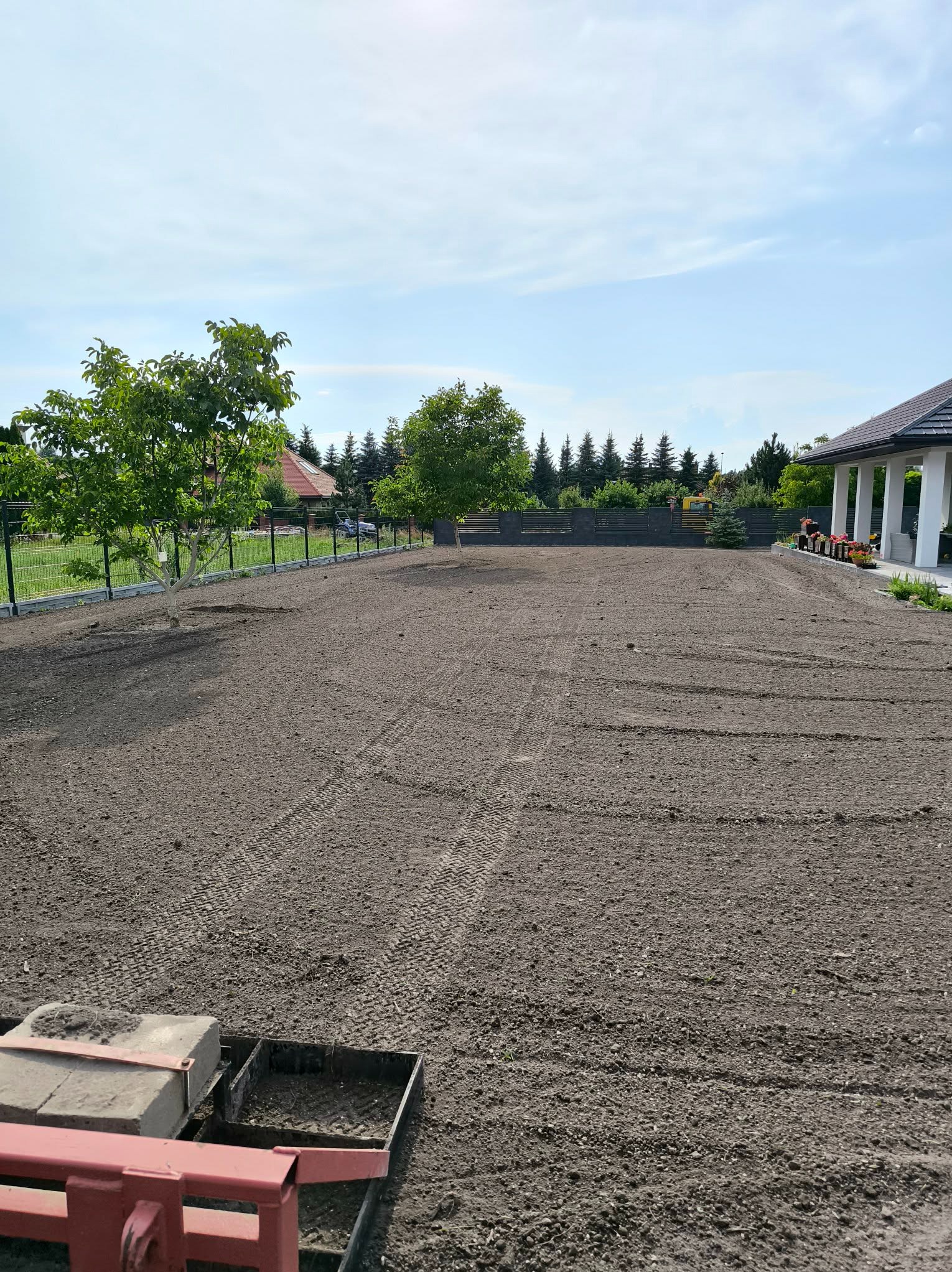 Przygotowany teren pod trawnik z widocznymi śladami po maszynach, drzewa w tle i fragment domu z kolumnami. Grunt wyrównany, gotowy do dalszych prac.