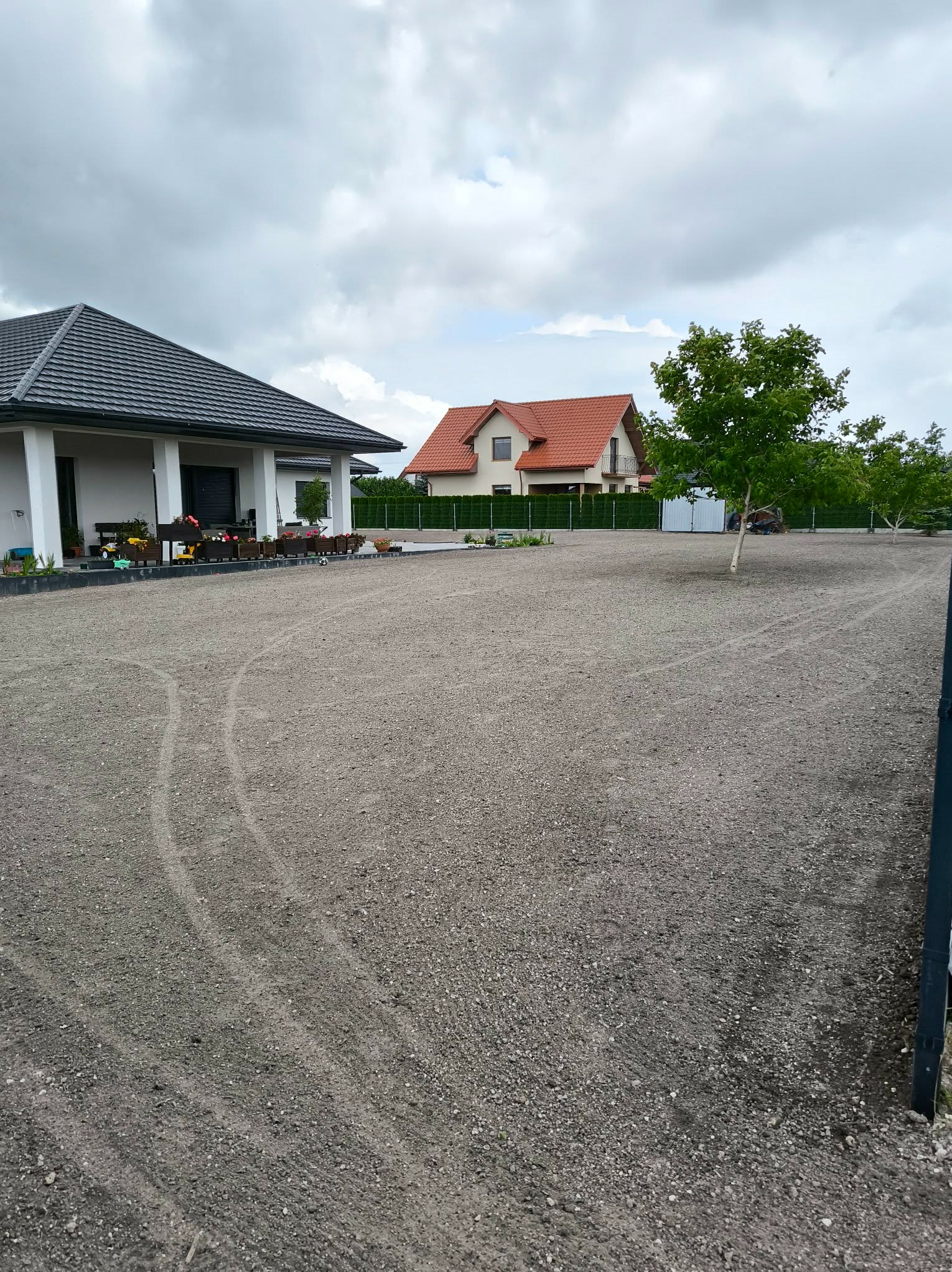 Przygotowany teren pod trawnik, widoczne ślady po maszynach, dom z szarą dachówką i rabaty kwiatowe w tle, pochmurne niebo.